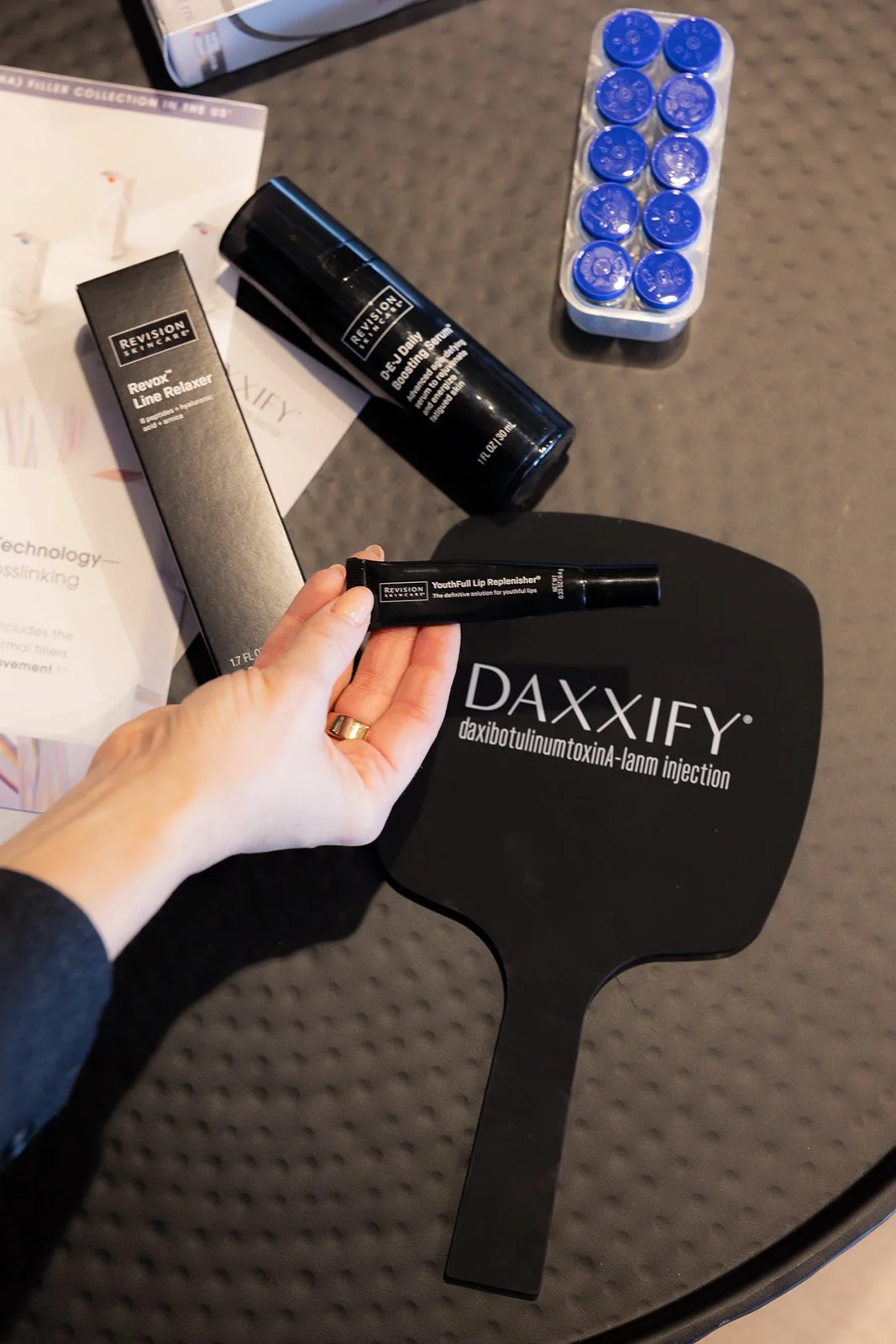 Assorted Botox and dermal filler products on a table, including Revance Revanesse, Revive D.E.P. Serum, Youthfull Lip Replenisher, and a display of blue capsules, with a DAXXIFY sign.