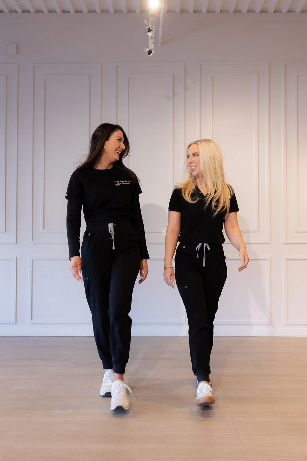 Two women dressed in black workout clothes walking and smiling indoors against a white paneled wall.