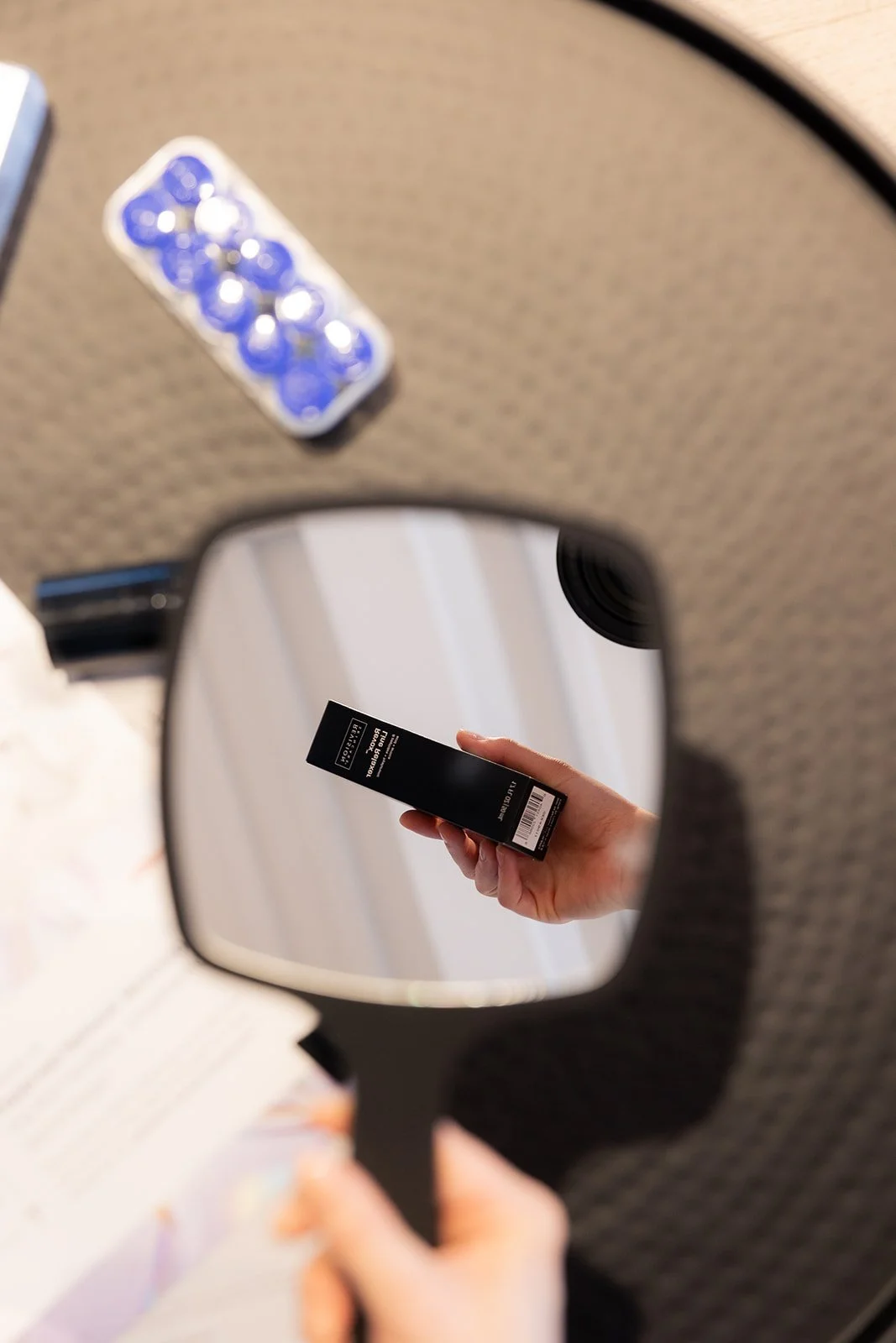 A person's hand holding a black boxed lipstick, visible through a mirror.