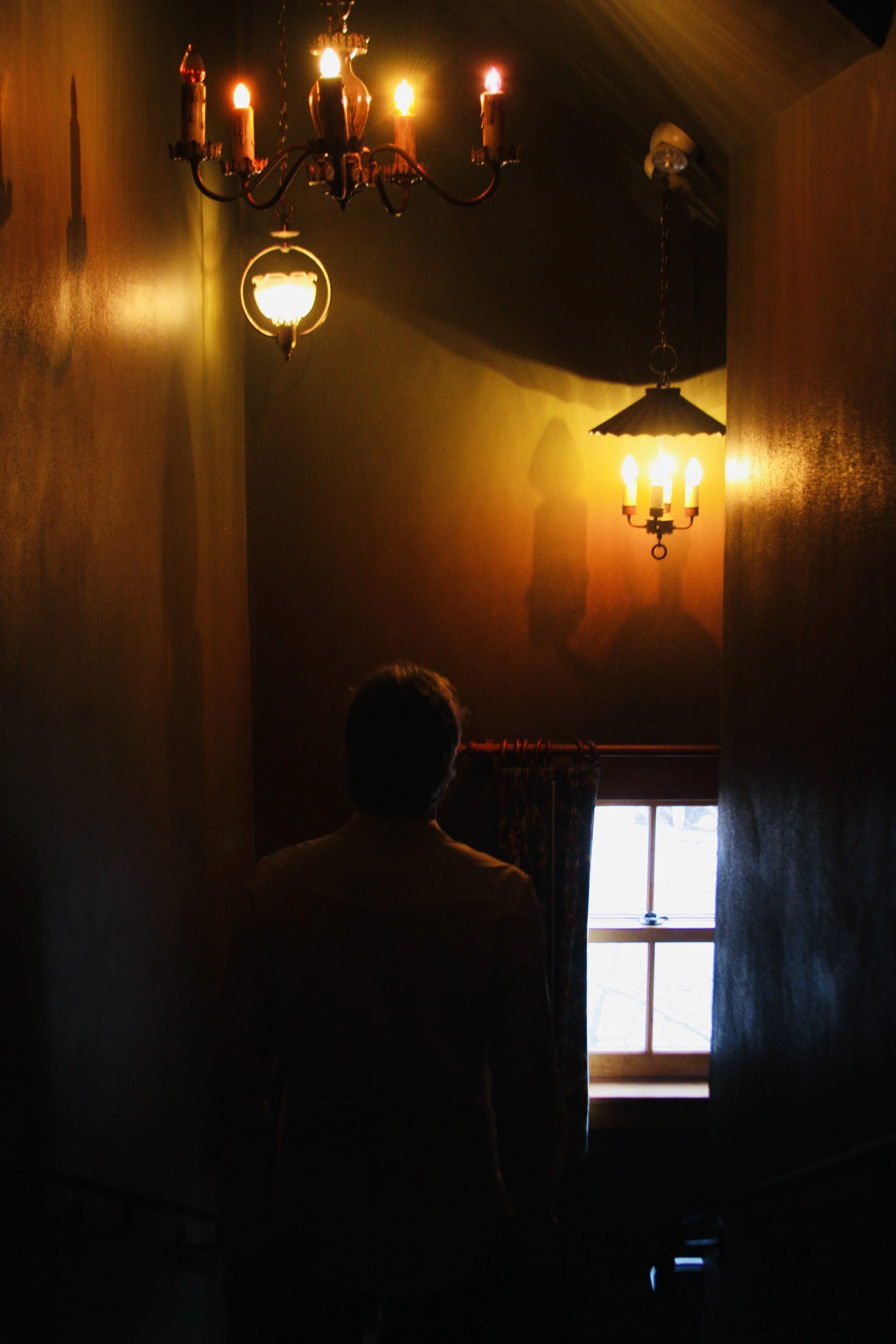 A person Justin Lavik standing at the top of a stairwell, looking out a window in a dimly lit room with warm lighting from chandeliers.