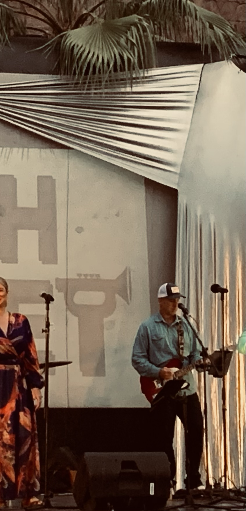 A man playing guitar and singing on stage with microphone, and a woman next to him, with a large screen behind them displaying abstract patterns. Part of a musical event or performance.