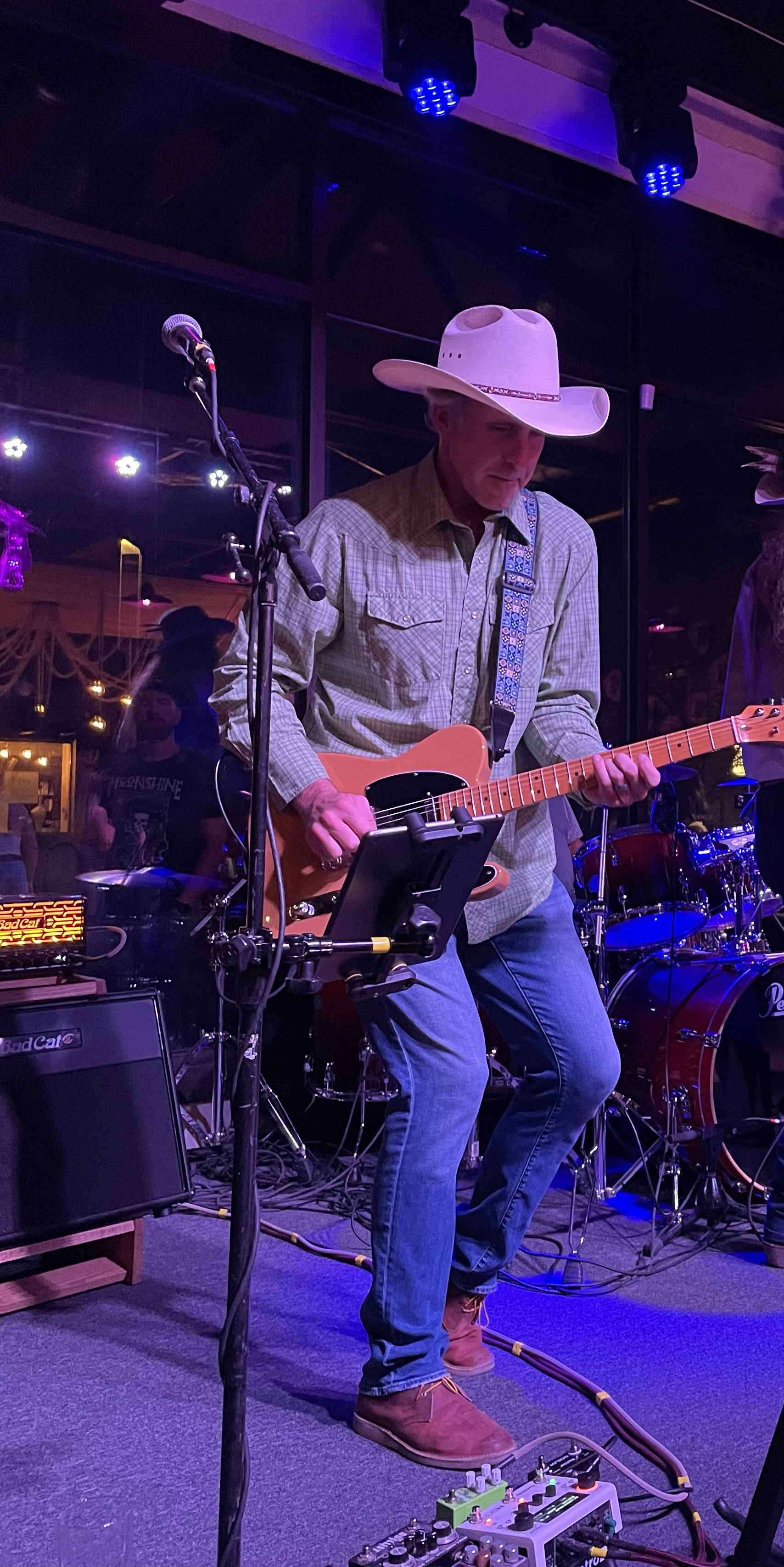A man wearing a cowboy hat, plaid shirt, and jeans playing an electric guitar on stage with background drums and colorful lights.