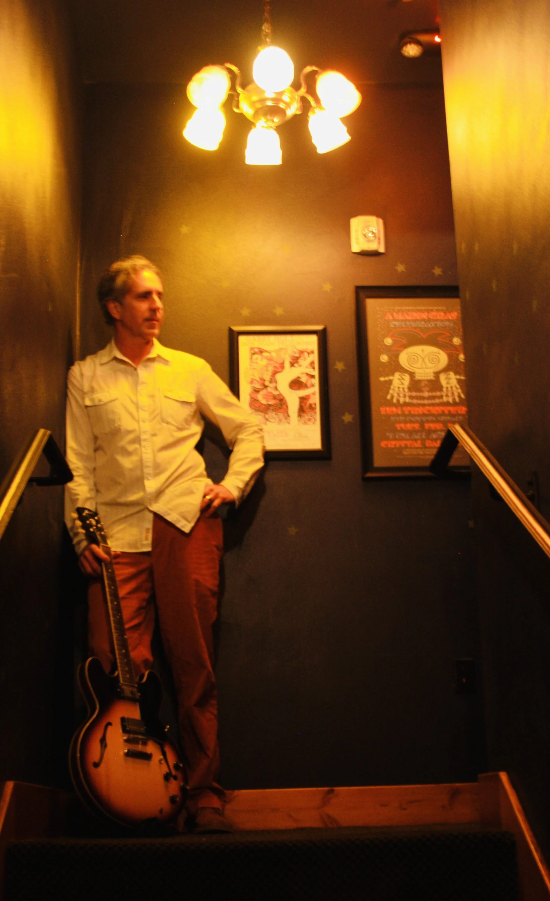 JUSTIN LAVIK A man standing on a small staircase landings holding an electric guitar, with a dark wall behind him decorated with framed artwork and a lit ceiling light above.