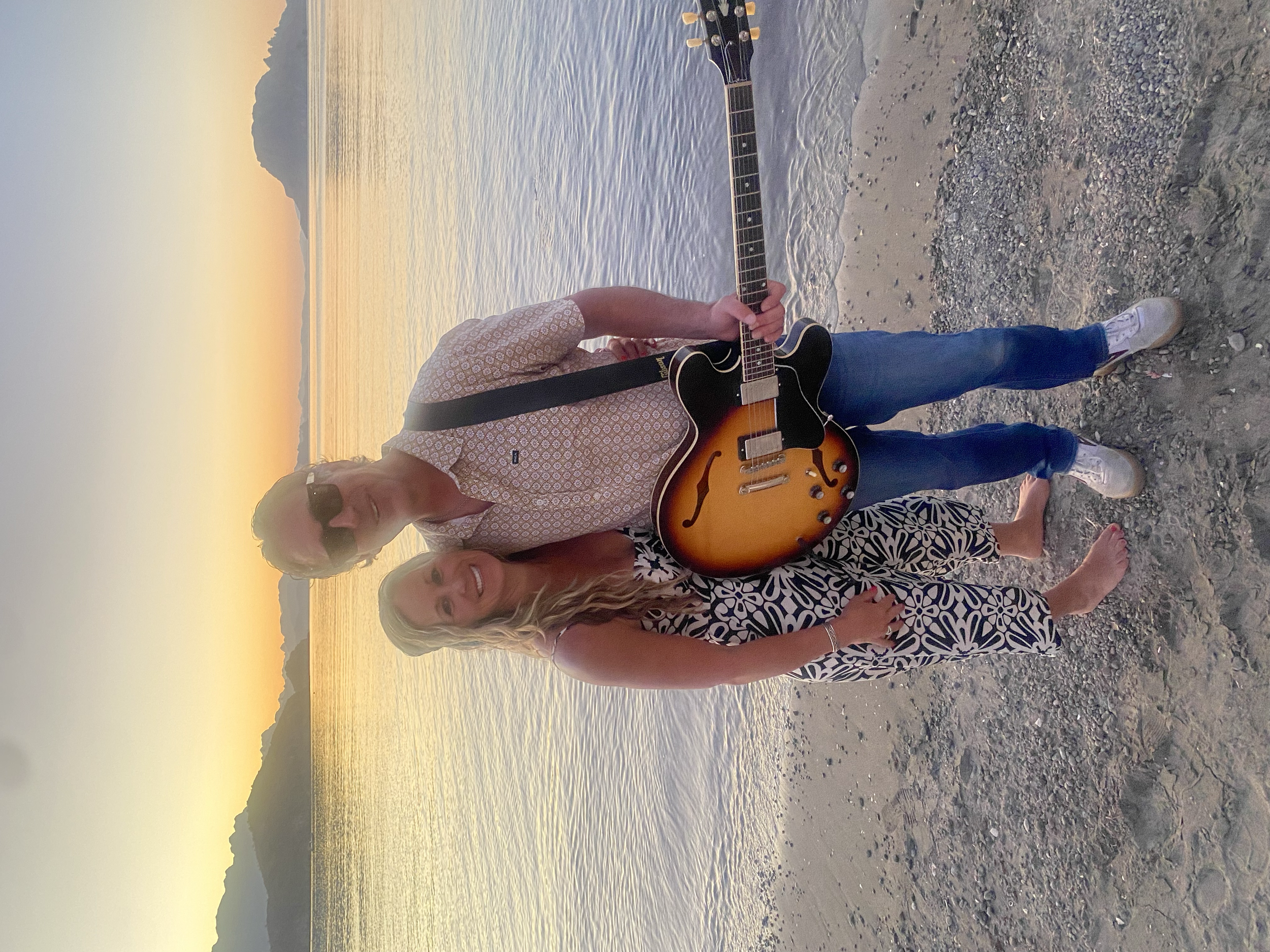 A man and a woman stand together on a rocky beach at sunset, with the man holding a guitar. The man wears sunglasses, a patterned short-sleeve shirt, and jeans, while the woman wears a patterned dress and is barefoot. Mountainous islands can be seen 