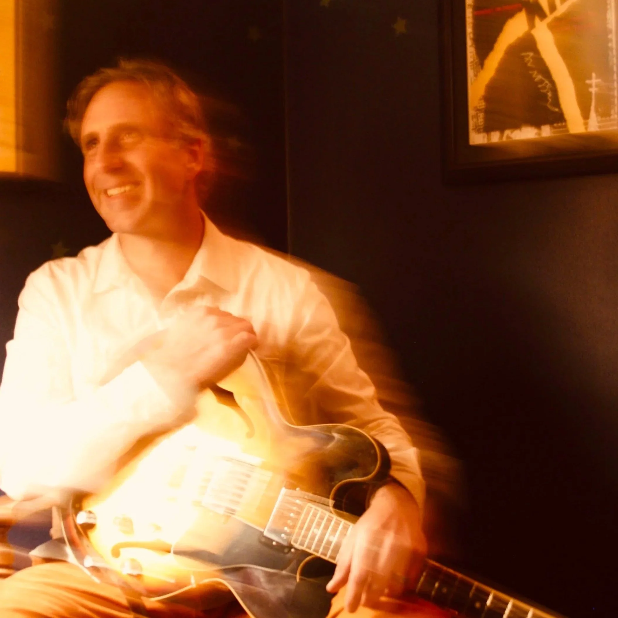 A man with a white shirt smiling and holding an electric guitar, with a motion-blur effect, in a dimly lit room with dark walls and a framed picture behind him.