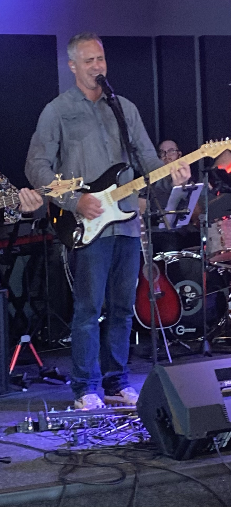 A man with gray hair playing an electric guitar and singing into a microphone on stage, surrounded by other musicians and musical equipment.