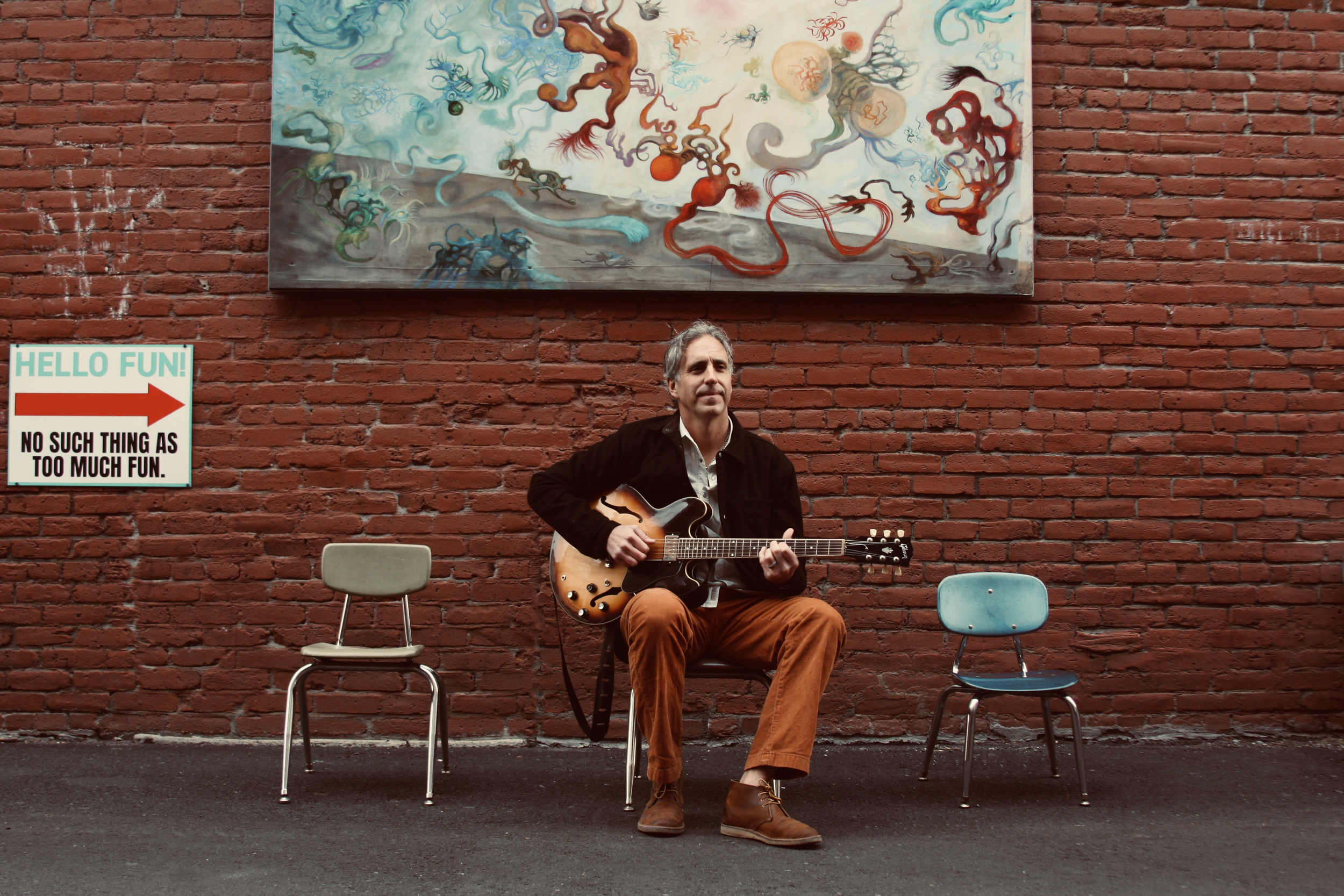 A man sitting on a chair playing an electric guitar in front of a brick wall with a colorful abstract painting above him. There are two empty chairs on each side and a sign to the left that reads 'HELLO FUN! NO SUCH THING AS TOO MUCH FUN' with a red arrow pointing right.