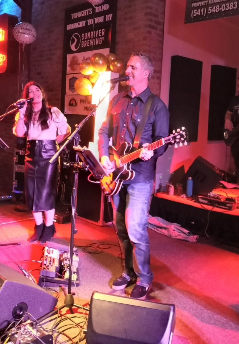 Musicians performing on stage at a live music venue. A woman with long dark hair in a white top and black leather skirt, and a man with short gray hair in a black shirt and jeans, playing an electric guitar. Bright stage lighting and rock band equipm