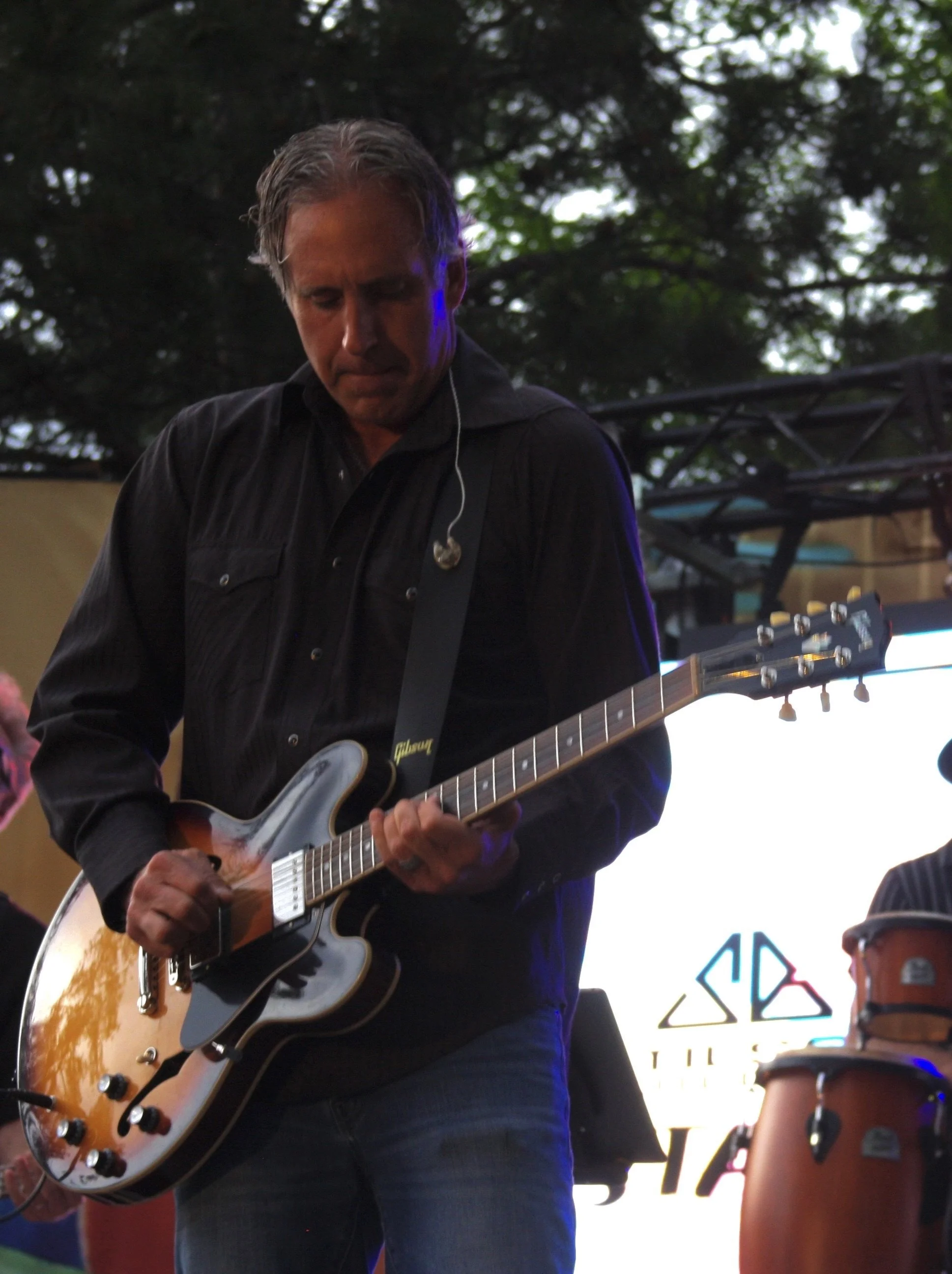 Musician playing an electric guitar on stage outdoors, with a background screen displaying a logo and trees overhead.