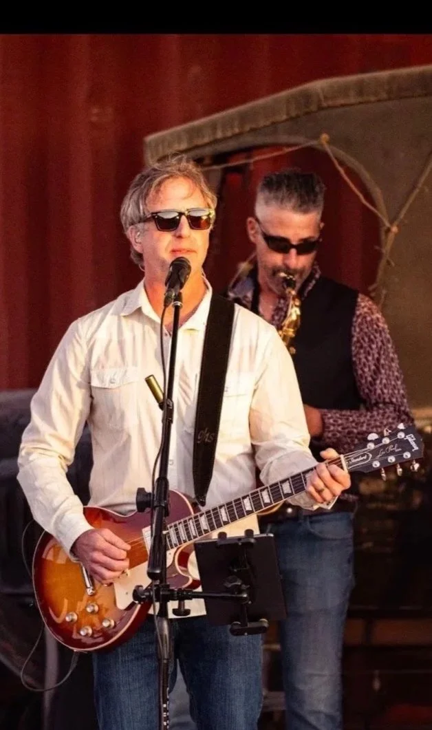 Two musicians performing on stage; one playing an electric guitar and singing into a microphone, the other playing a saxophone, both wearing sunglasses.