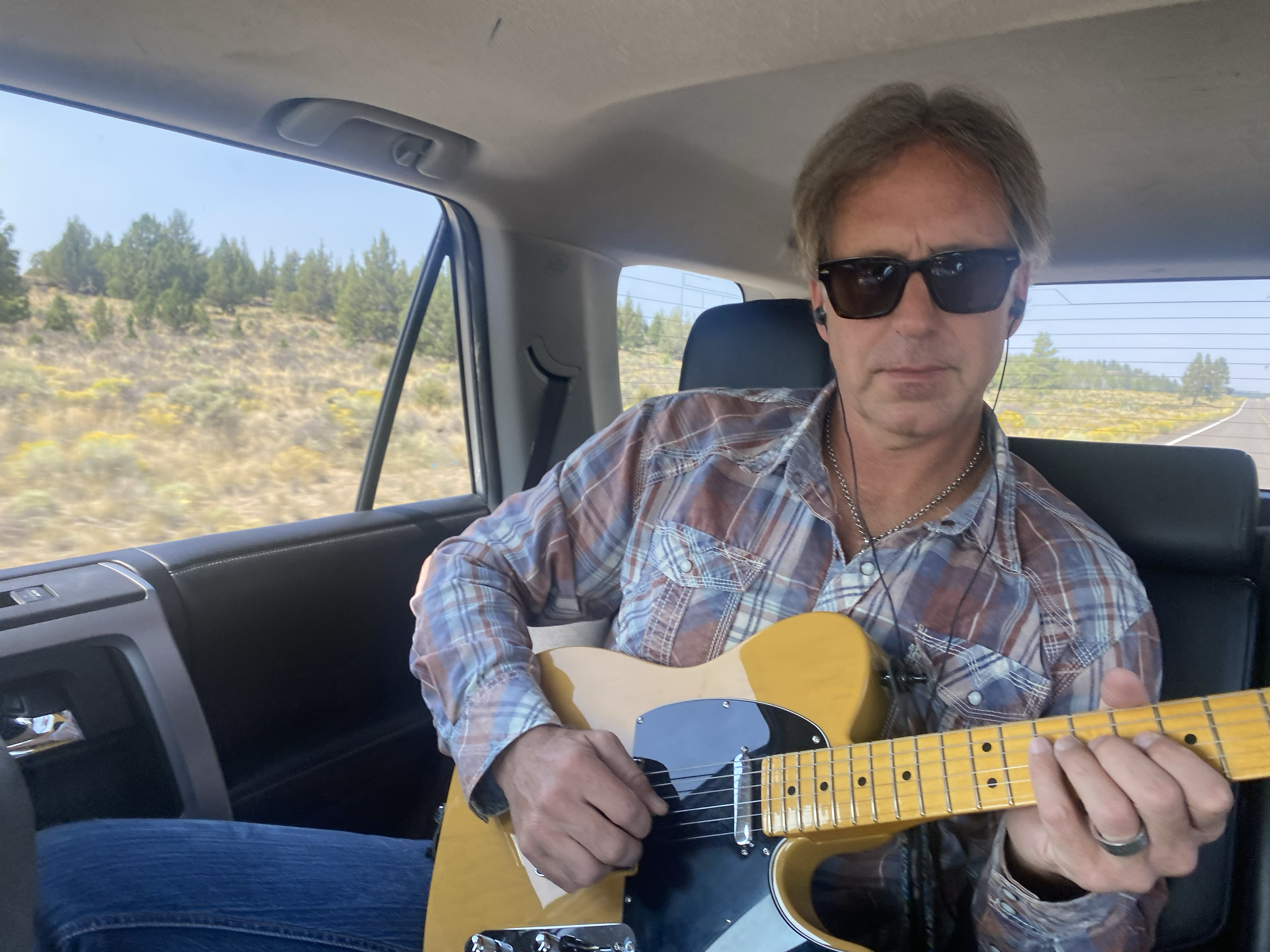 A man sitting in a vehicle, wearing sunglasses, plaid shirt, and earphones, playing an electric guitar with a landscape of trees and blue sky visible through the window.