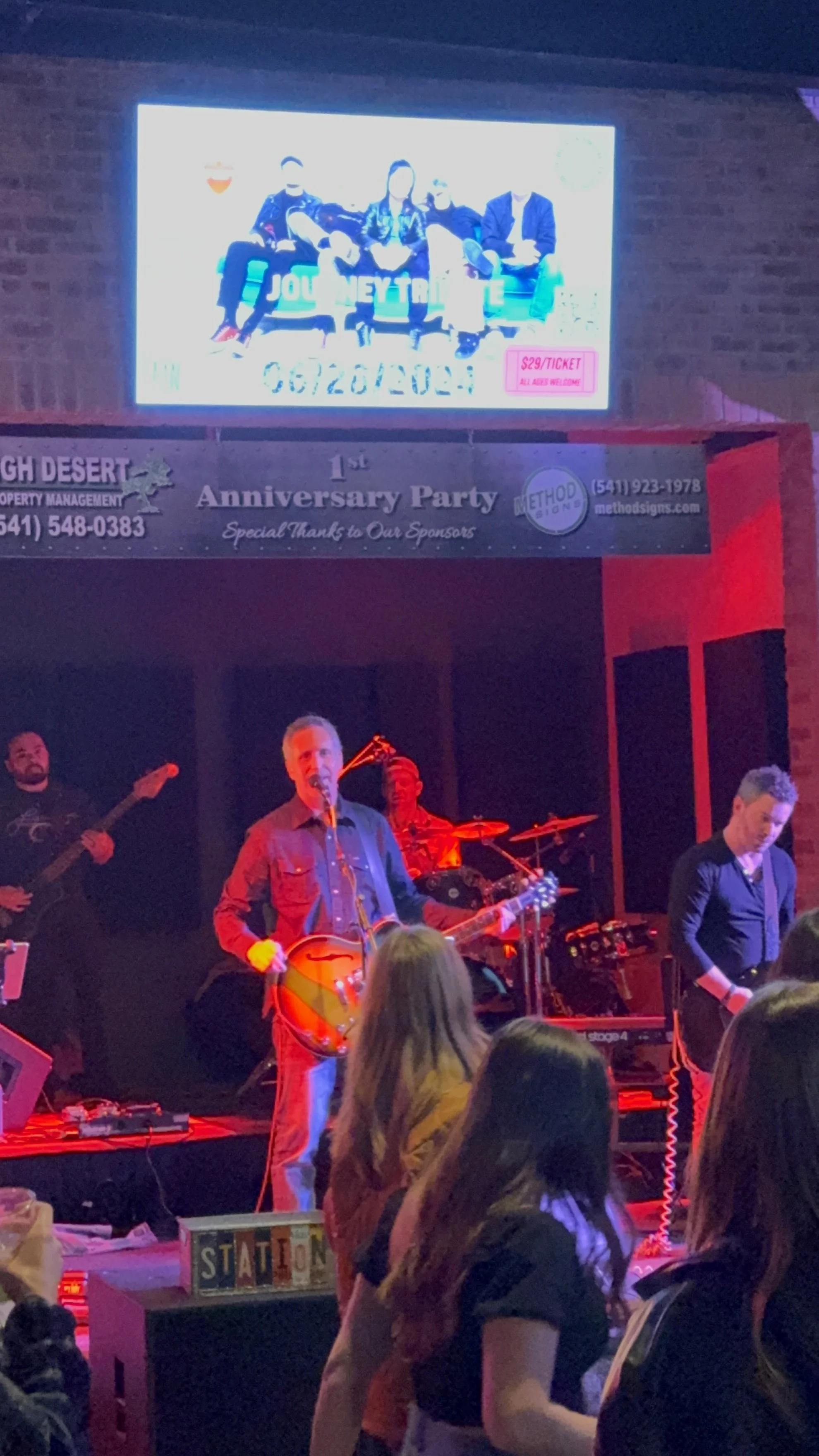 A band performing on stage at an event with an audience watching, with a large screen above showing people sitting on a bench and ticket pricing information.