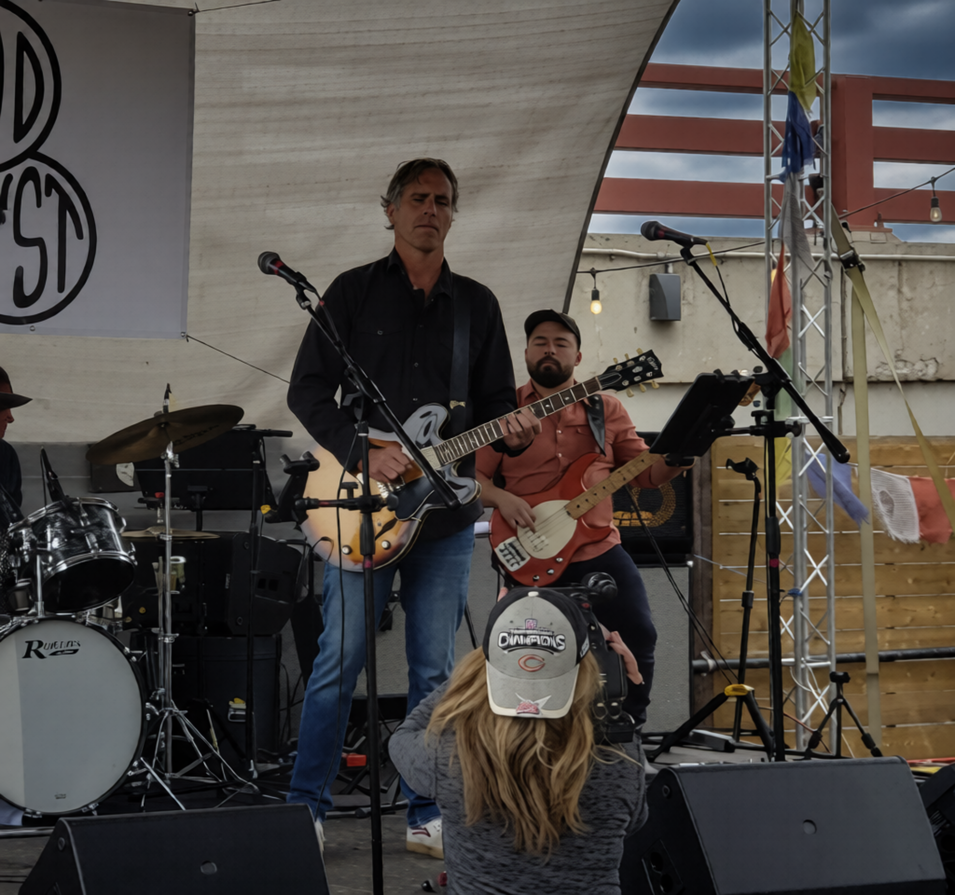 A band performing on an outdoor stage with two guitarists, drums, and a person in the foreground taking photos. The sky is cloudy, and there is a banner with a partial logo on the left side. A woman with blonde hair wearing a baseball cap is in the f