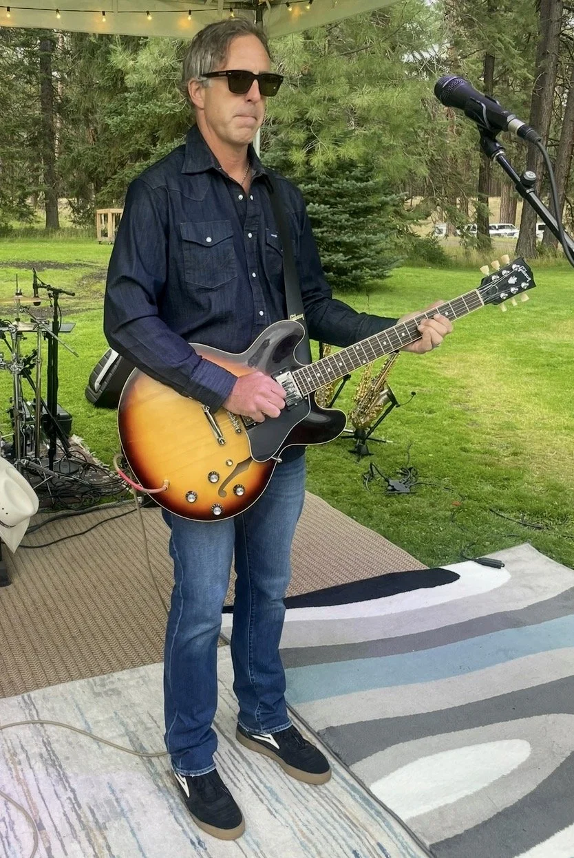 A man wearing sunglasses, a black shirt, and jeans plays an electric guitar on an outdoor stage with a microphone nearby. There is musical equipment and trees in the background.