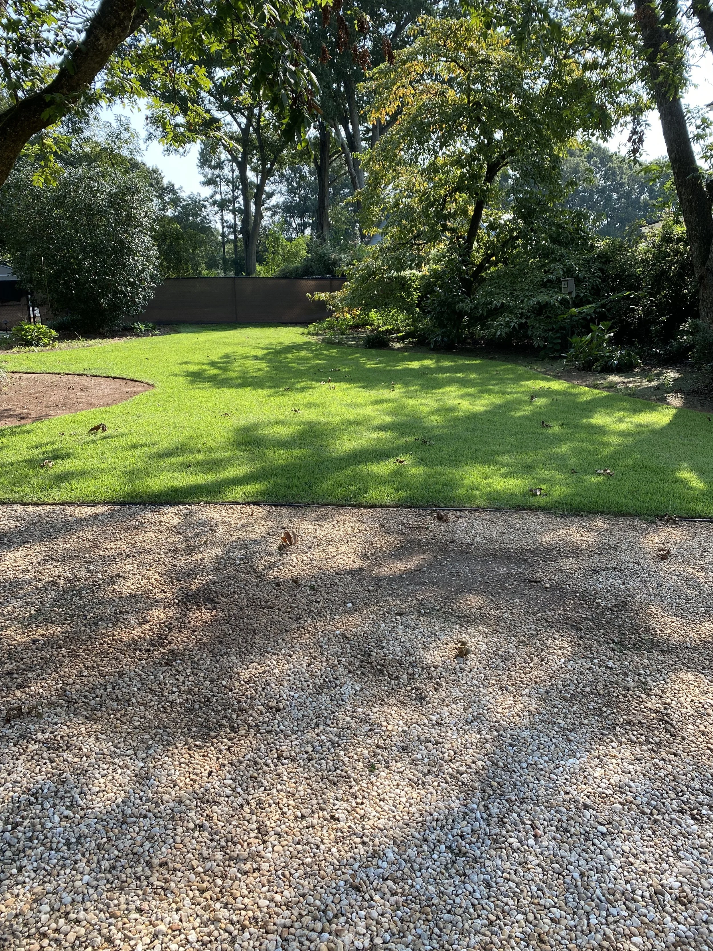 A backyard with a gravel patio in the foreground and a lush green lawn surrounded by trees and bushes in the background, with sunlight casting shadows.