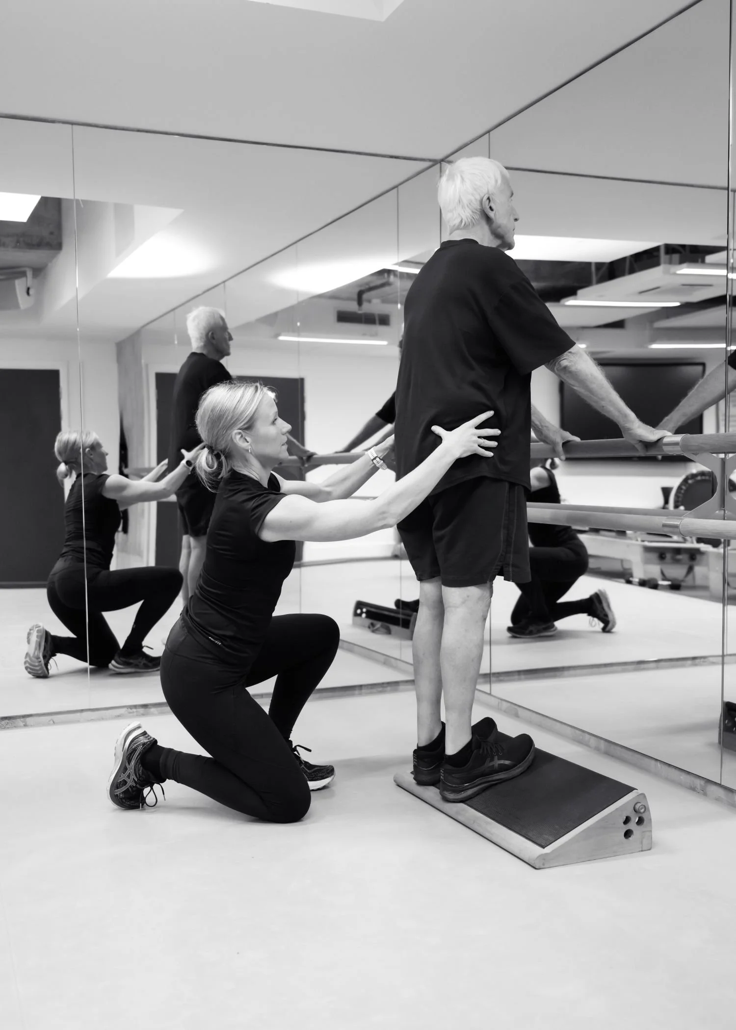 A physical therapist assisting an elderly man during a rehabilitation exercise in a gym with mirrored walls, with other individuals in the background participating in exercises.