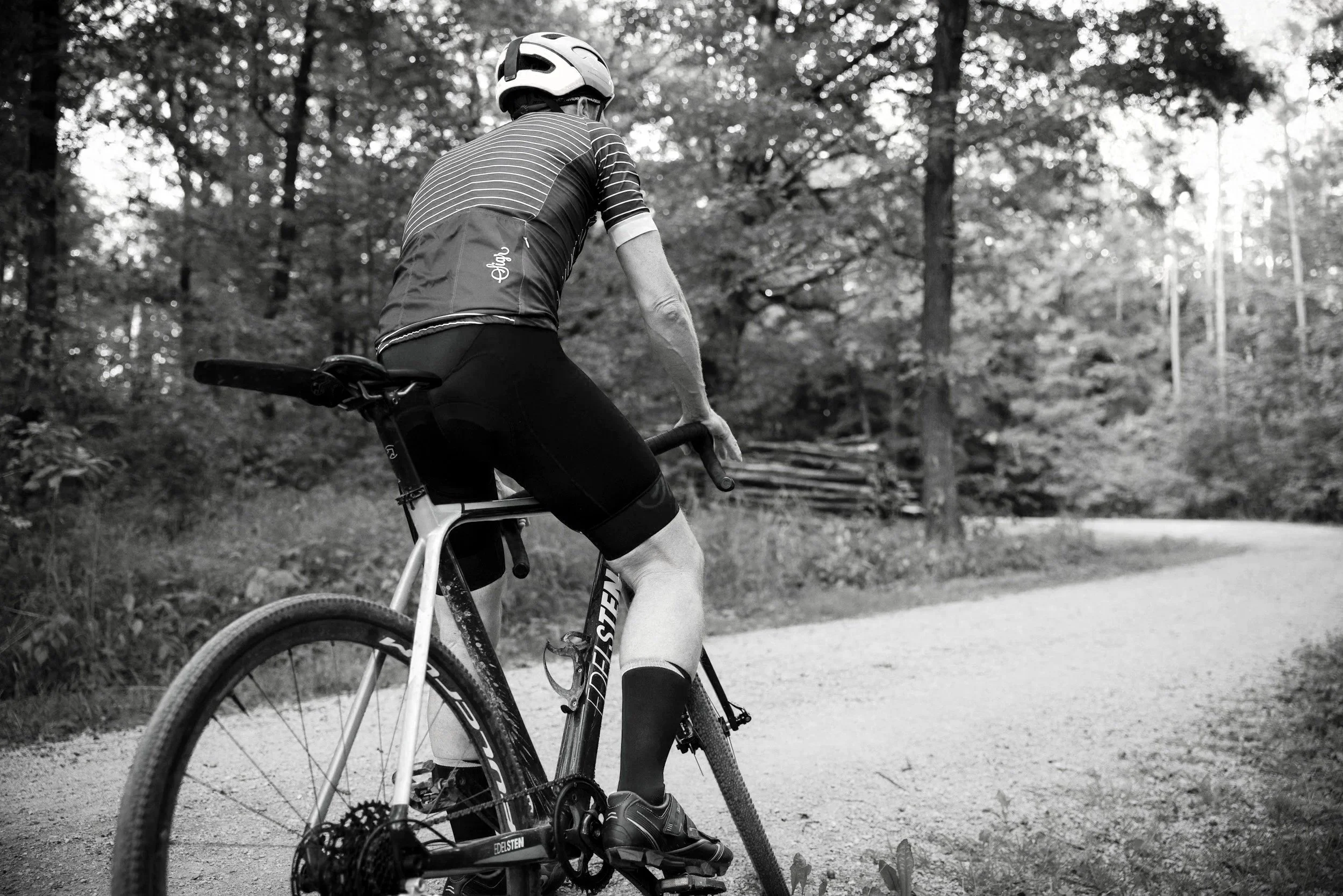 A person riding a mountain bike on a trail in a wooded area, wearing a helmet and cycling gear, captured from the back in black and white.