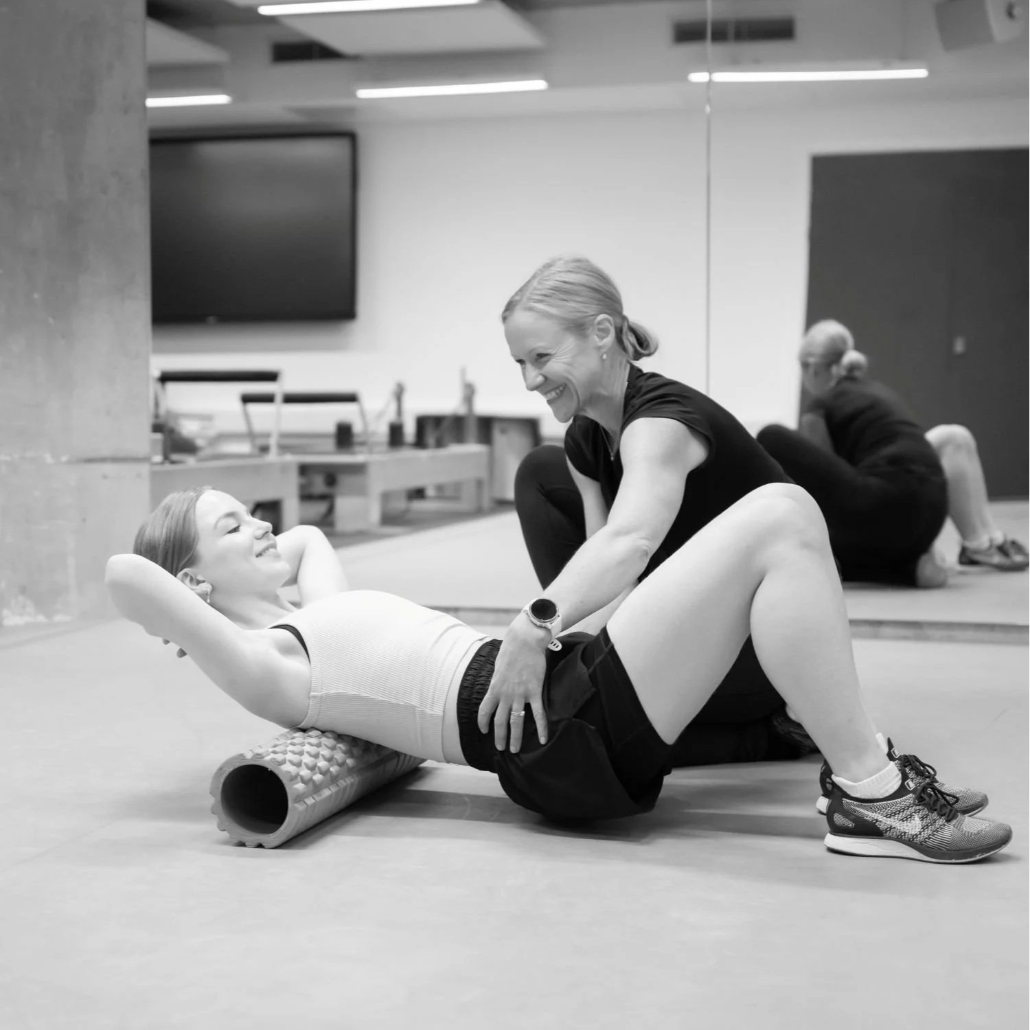 A woman in a gym setting with a smile lying on a foam roller with her knees bent, engaging in physiotherapy exercisess with a Anna Turgoose.