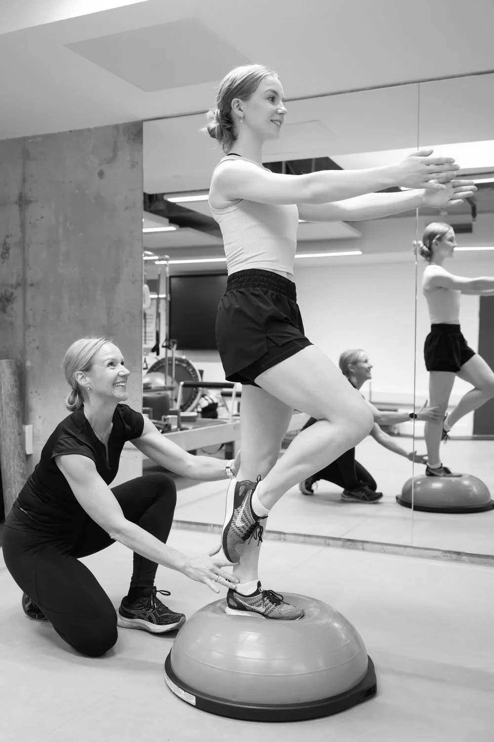 A young woman doing balance exercises on a Bosu ball with assistance from an instructor in a gym, reflected in a mirror.