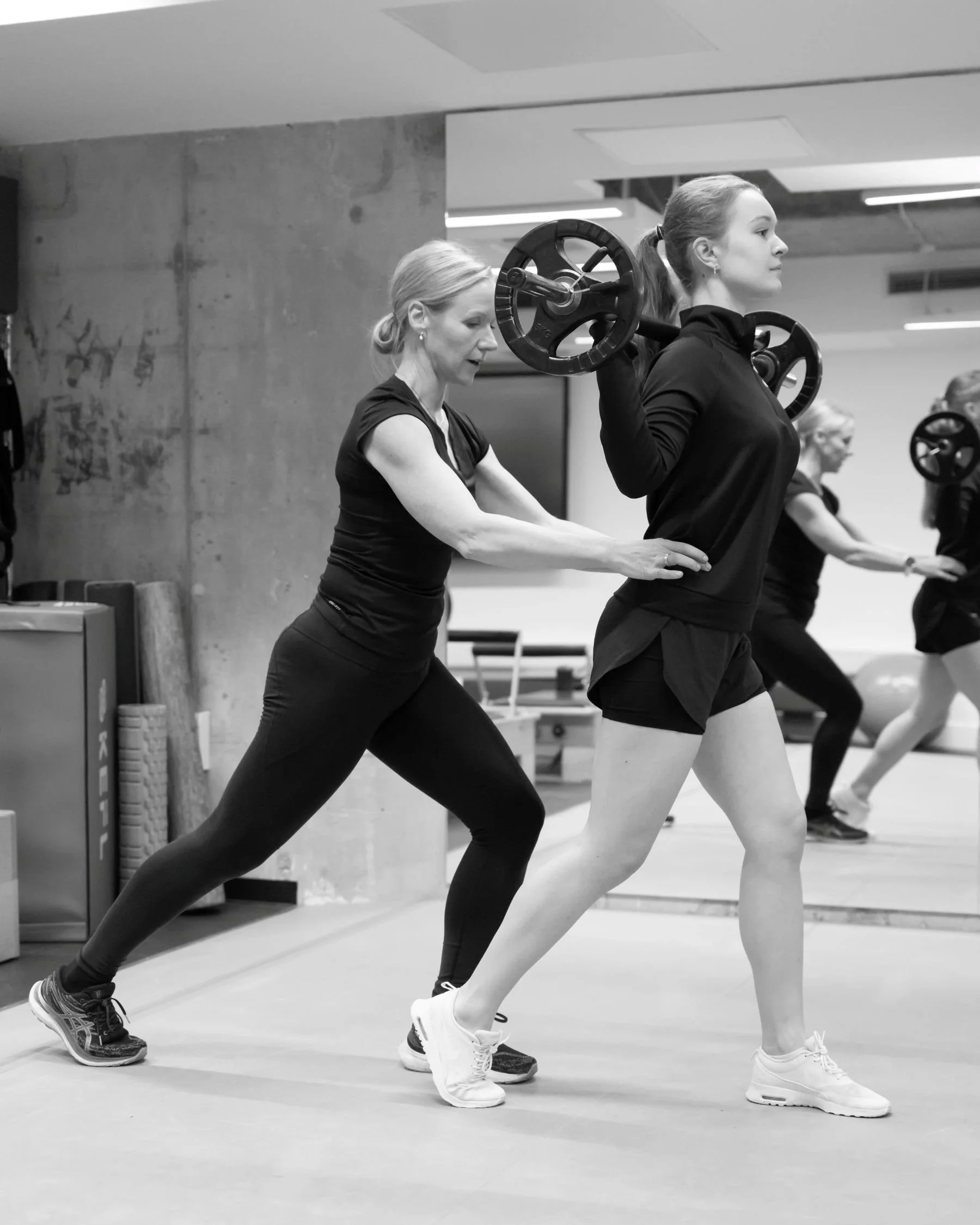 A woman is assisting another woman during a weightlifting exercise at the gym, with both wearing athletic clothes.