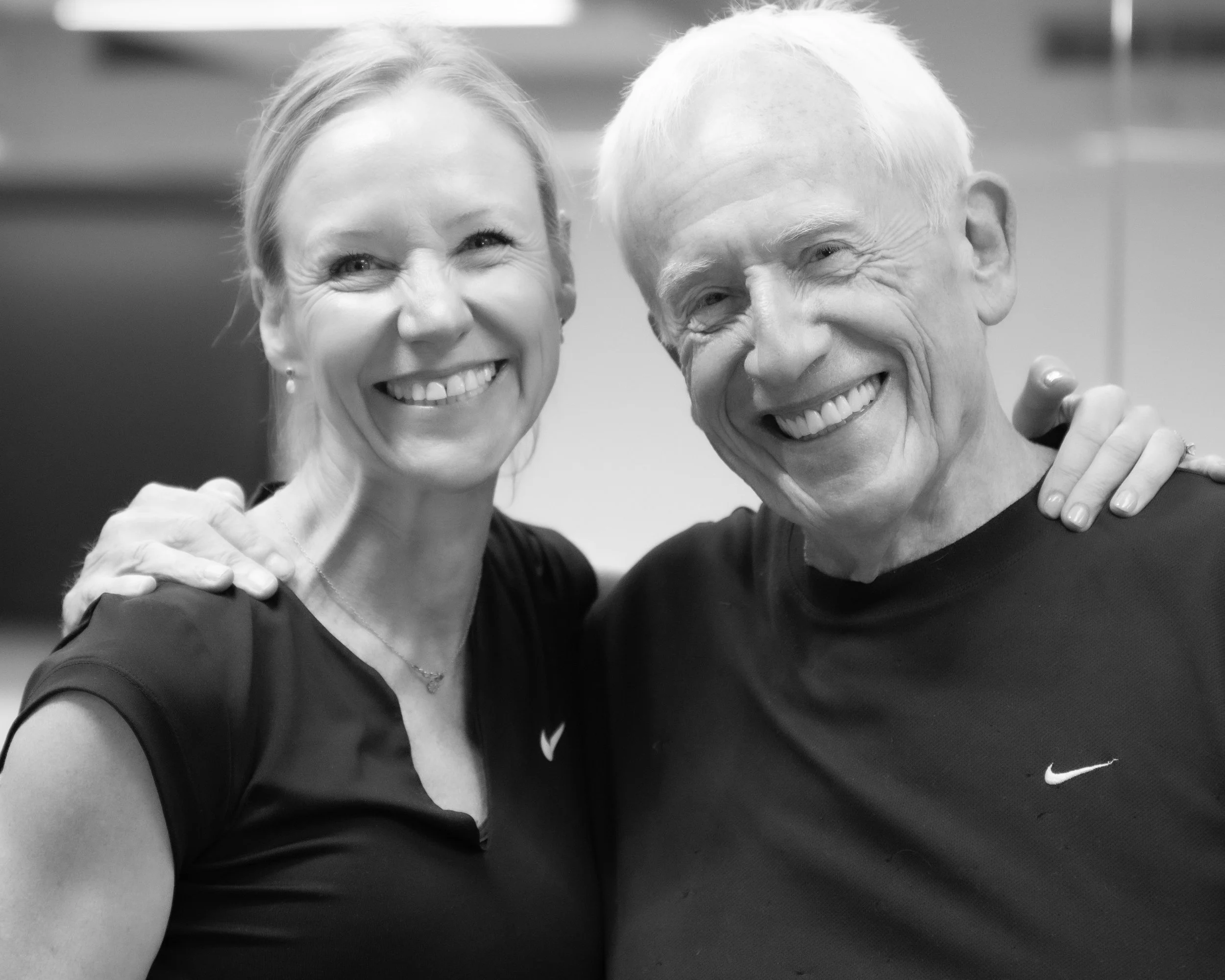 A smiling woman and man in athletic wear, both with their arms around each other, in a gym or fitness setting.