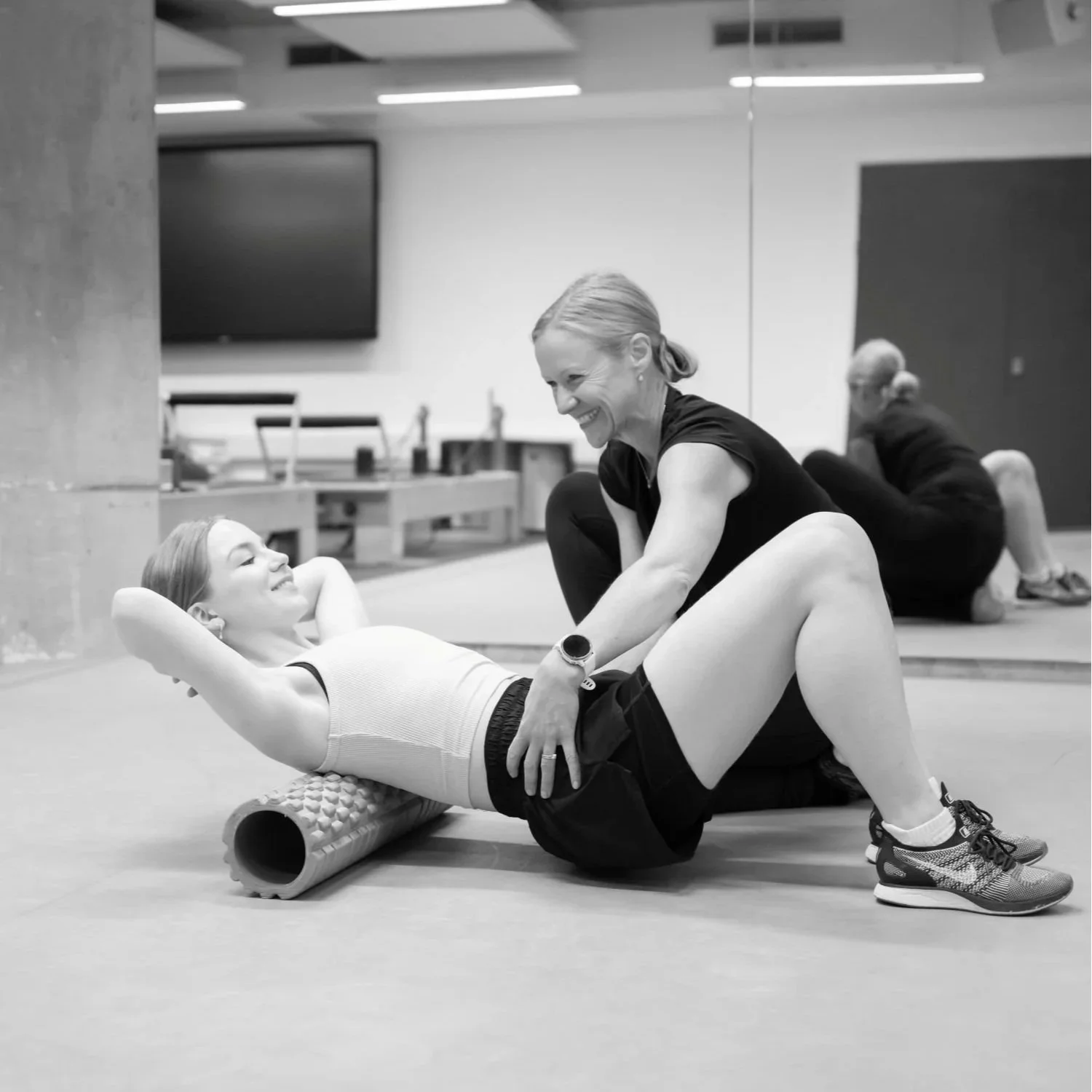 Two women engaging in a fitness class, one lying on a foam roller with her hands behind her head and smiling, the other kneeling beside her, smiling and assisting. In the background, another woman is performing a stretch or exercise on the floor.