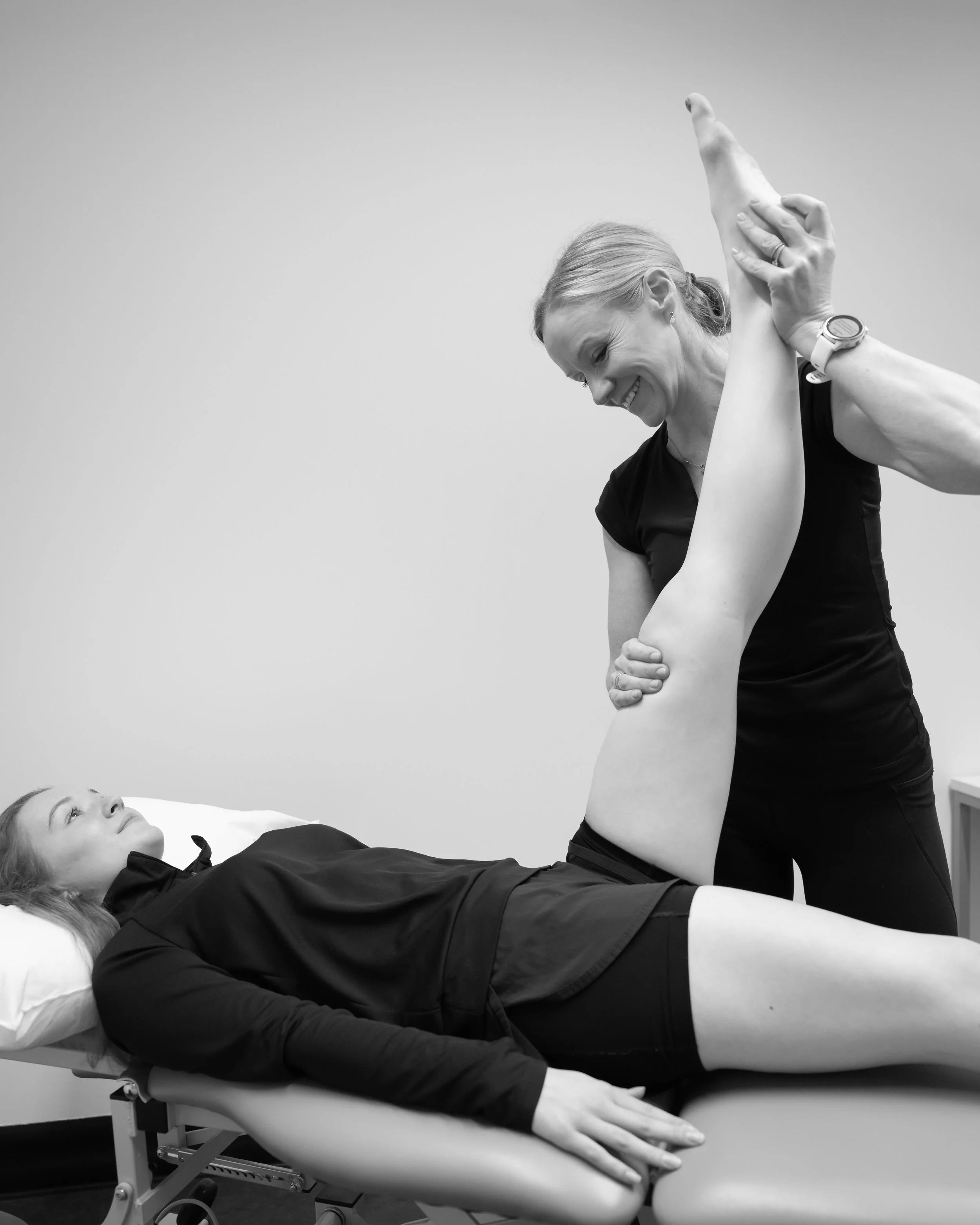A woman in a black outfit is performing a stretching or physical therapy exercise on a woman lying on an examination table, in a clinical or fitness setting.