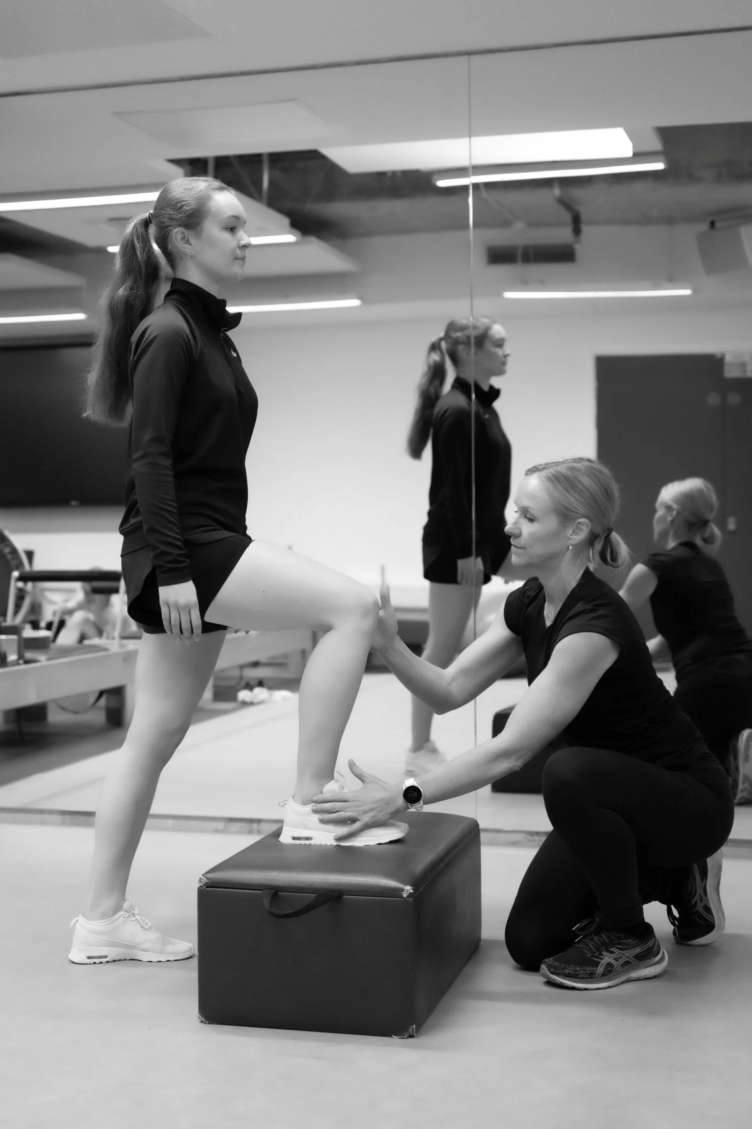 A fitness trainer helping a woman with her knee during a workout in a gym, with a mirror and other women exercising in the background.