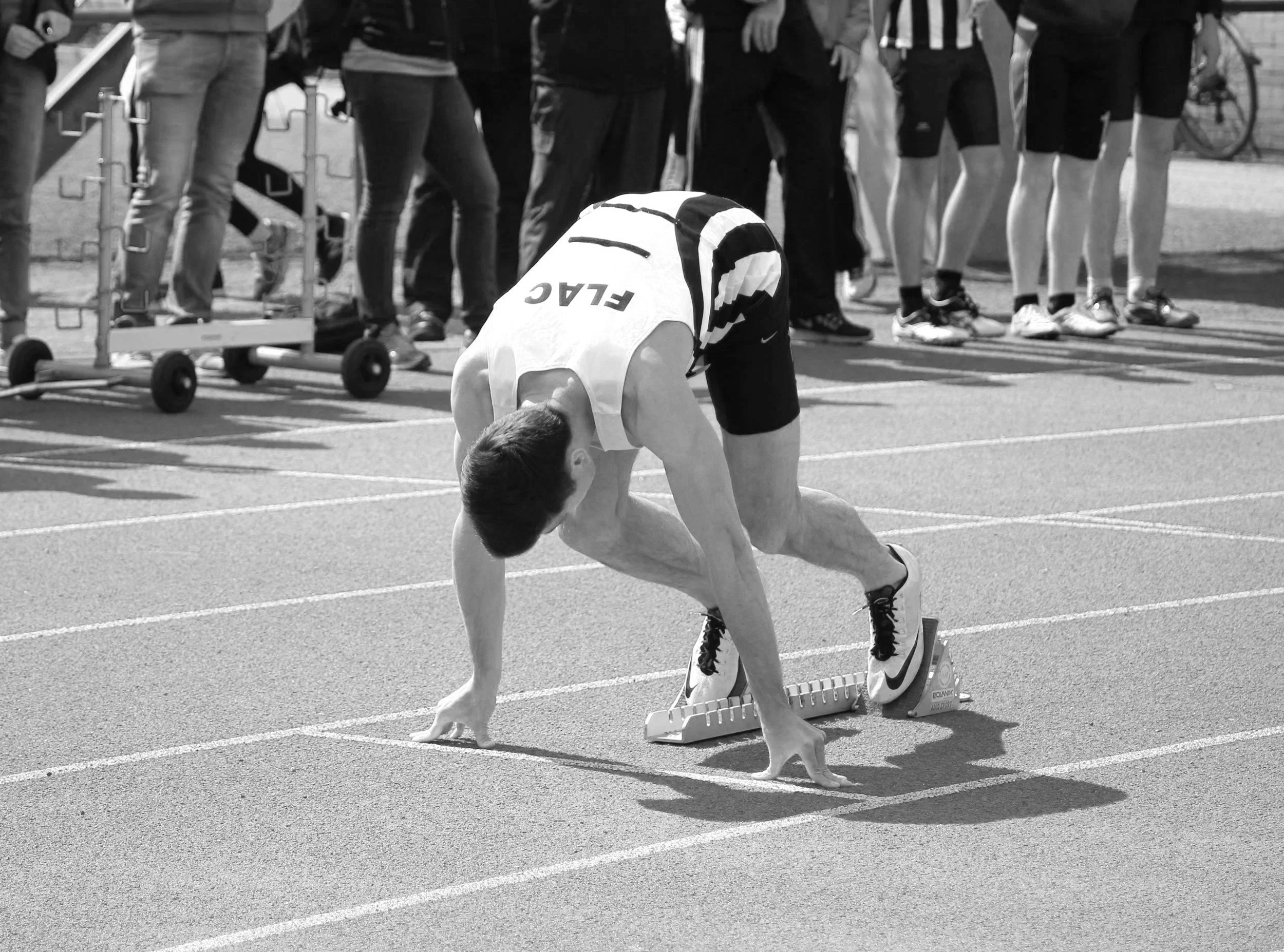 A male sprinter in starting position on a track, preparing for a race, with spectators and athletes standing behind him.