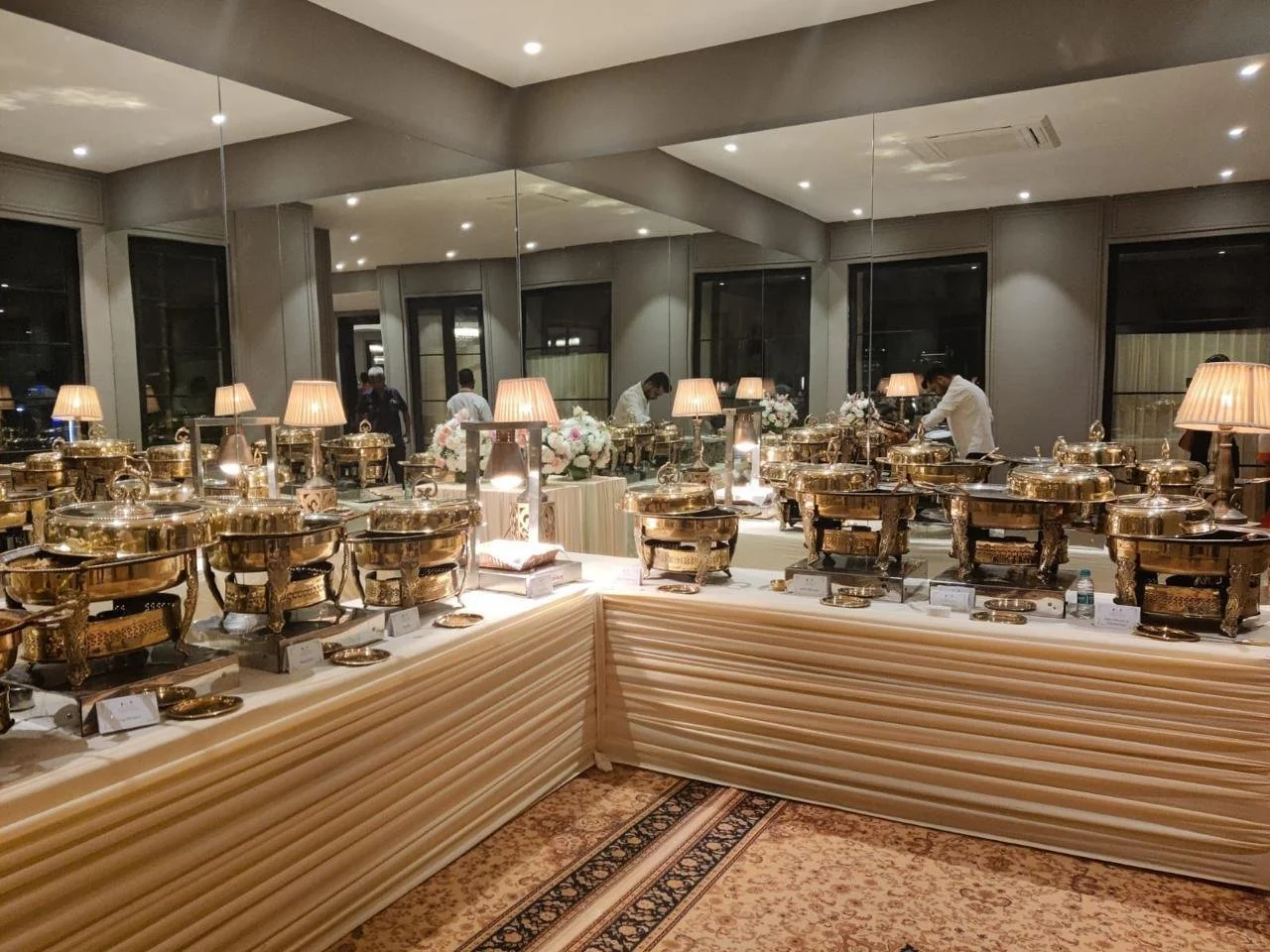 A buffet table with multiple gold chafing dishes and lamps, decorated with flowers, set in a banquet hall or event space.
