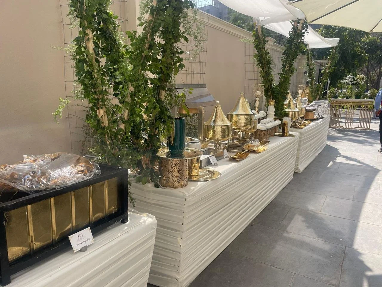 Decorative buffet table with gold and metallic serving ware, crockery, and plants outdoors under umbrellas.