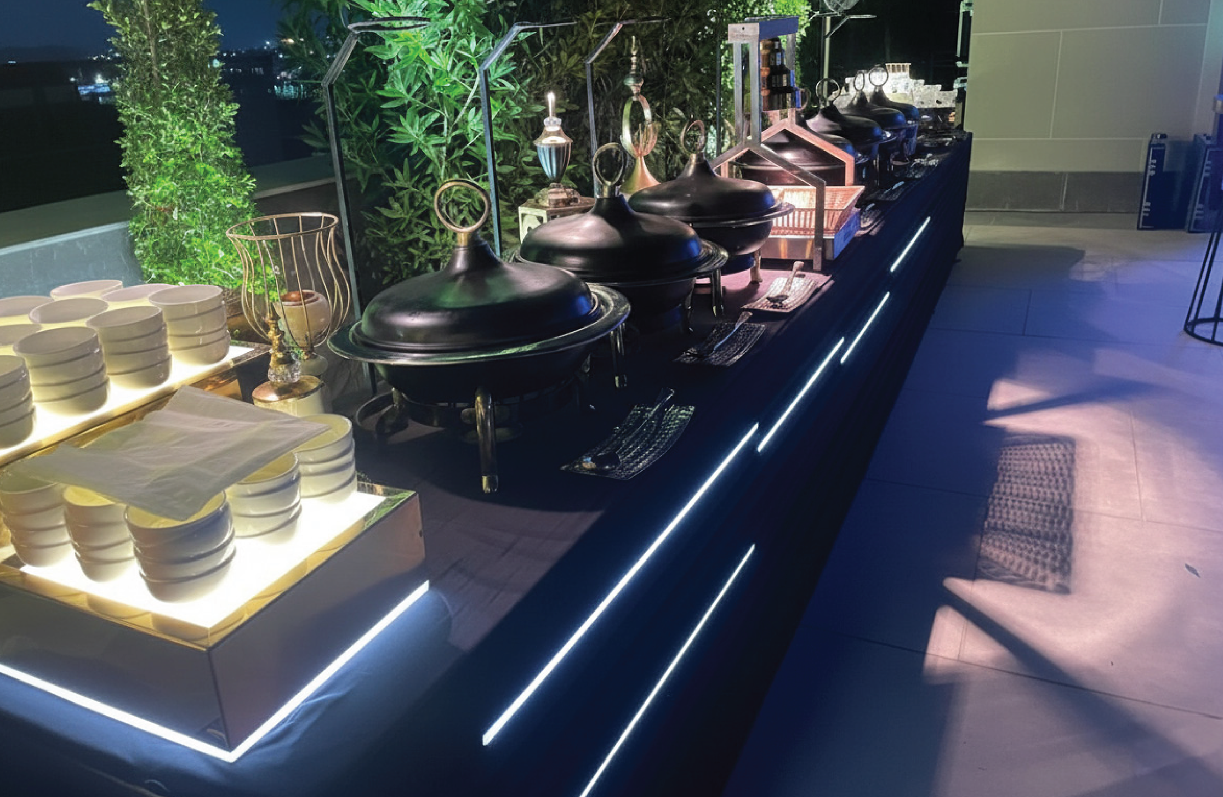 Buffet setup with stacked white bowls, three chafing dishes, and various decorative items on a black counter with blue lighting, against a backdrop of green plants and a night sky.