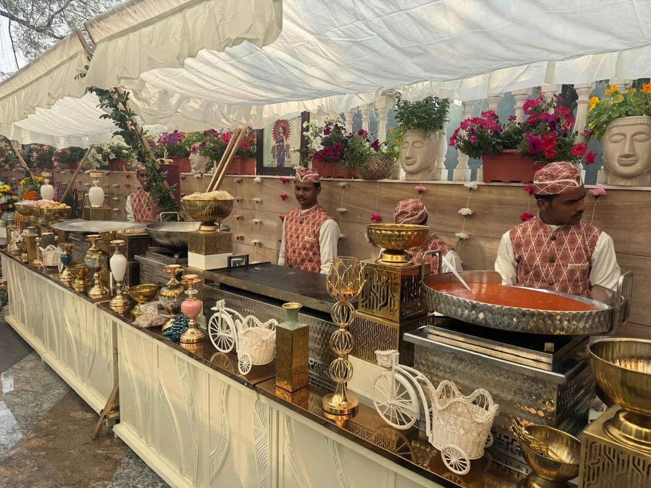Food stall at an event with four men in traditional attire standing behind a decorated counter with gold and white accessories, flowers, and religious images in the background.