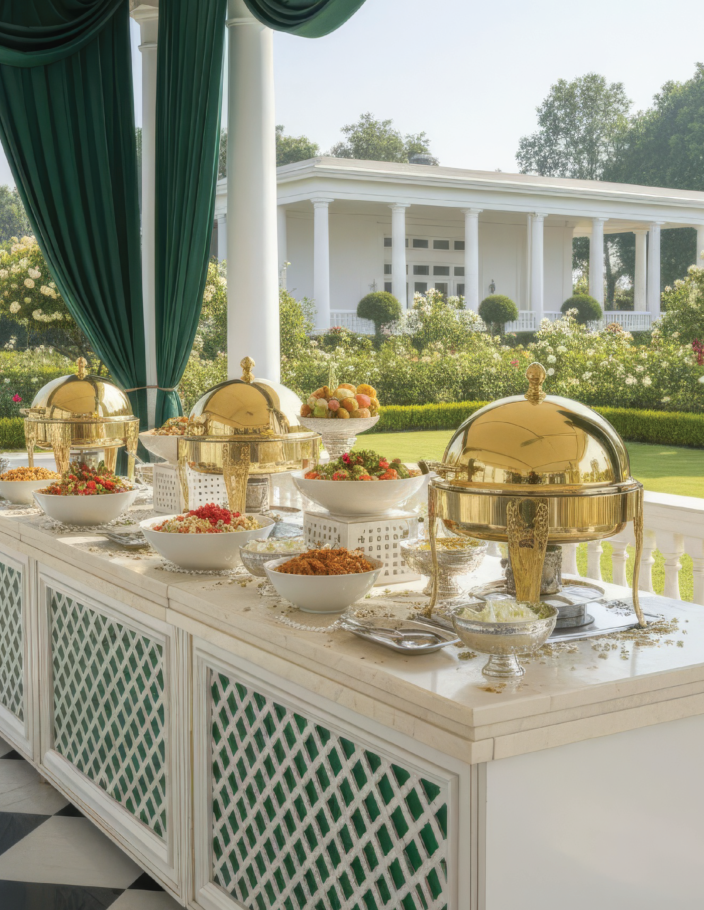Elegant outdoor buffet with chafing dishes and bowls of salad on a white table, overlooking a lush garden and a white house with columns.