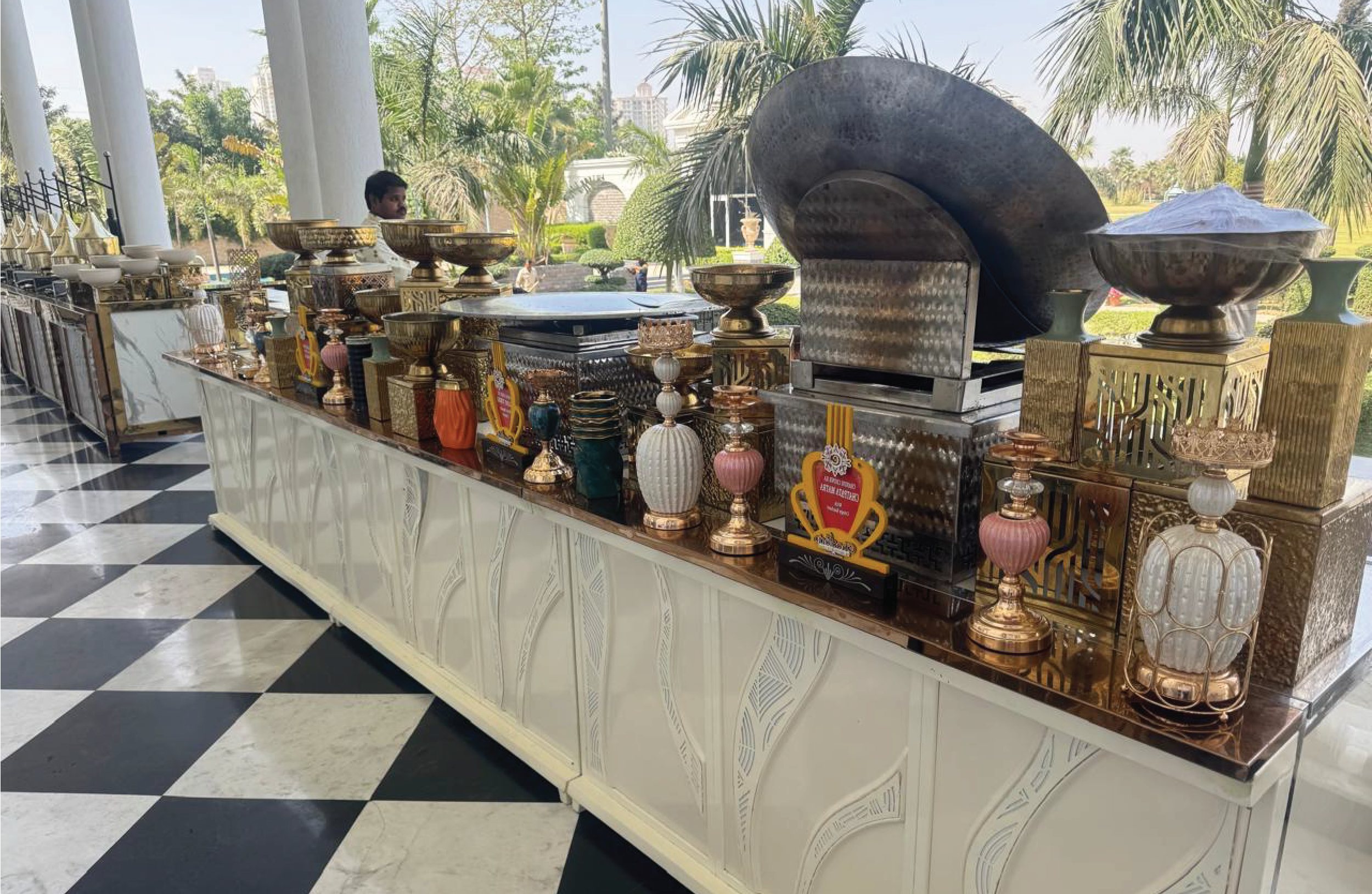 A buffet setup with various decorative vases, bowls, and trophies on a white table with a marble top, with a large outdoor lounge area and palm trees in the background.