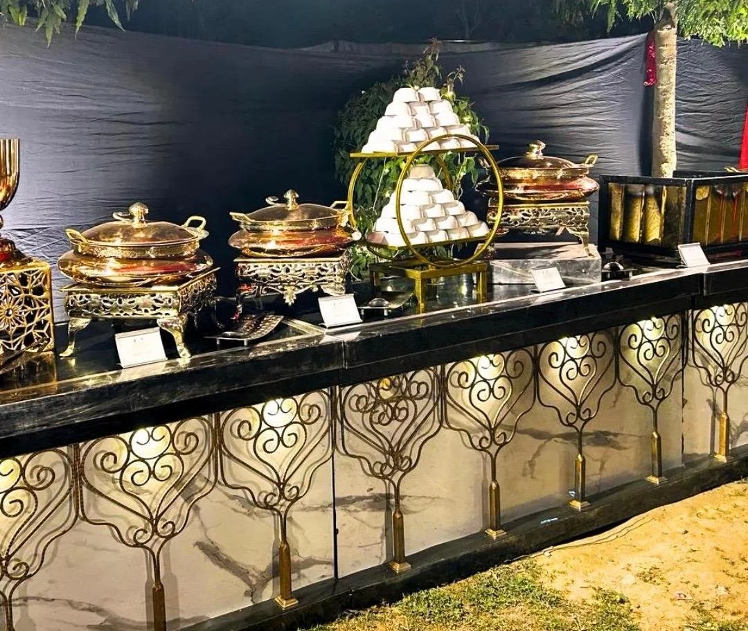 Buffet table with gold chafing dishes, a decorative frame with white cupcakes, and a black marble countertop in a dimly lit setting.