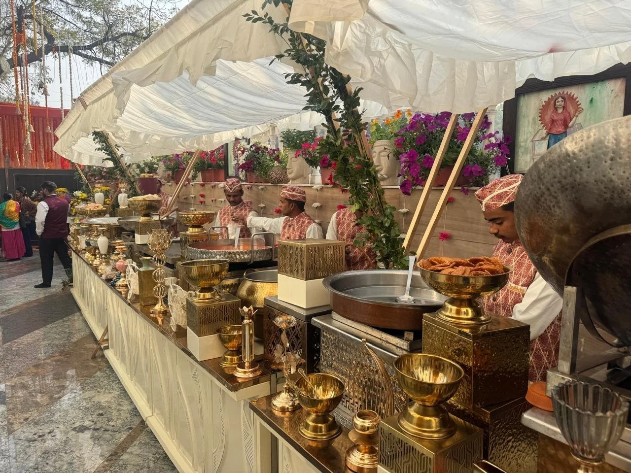A buffet setup at a celebration with various gold and silver decorative items, bowls, and food containers. Men in traditional Indian attire are serving or preparing food behind the counter, with flowers and greenery decorating the background.