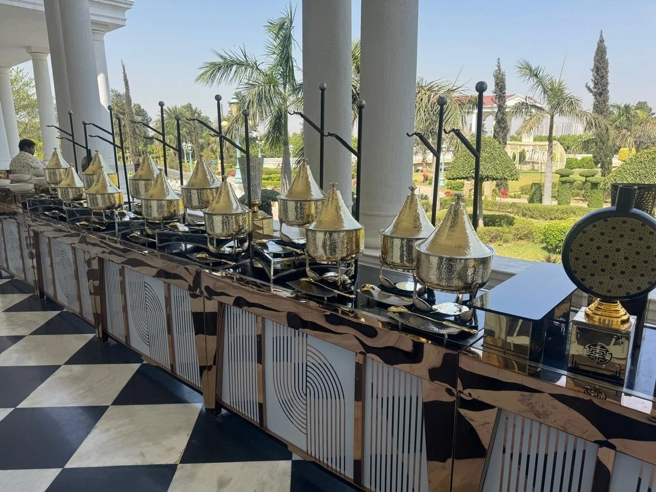 A buffet setup with multiple brass chafing dishes and serving utensils on a mirrored counter, next to a window overlooking a garden with palm trees and manicured bushes.