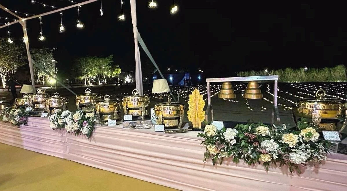 Nighttime outdoor event setup with a long table decorated with pink fabric and floral arrangements, featuring multiple golden bowls or chafing dishes, a small lamp, and other small items, with string lights overhead and trees in the background.