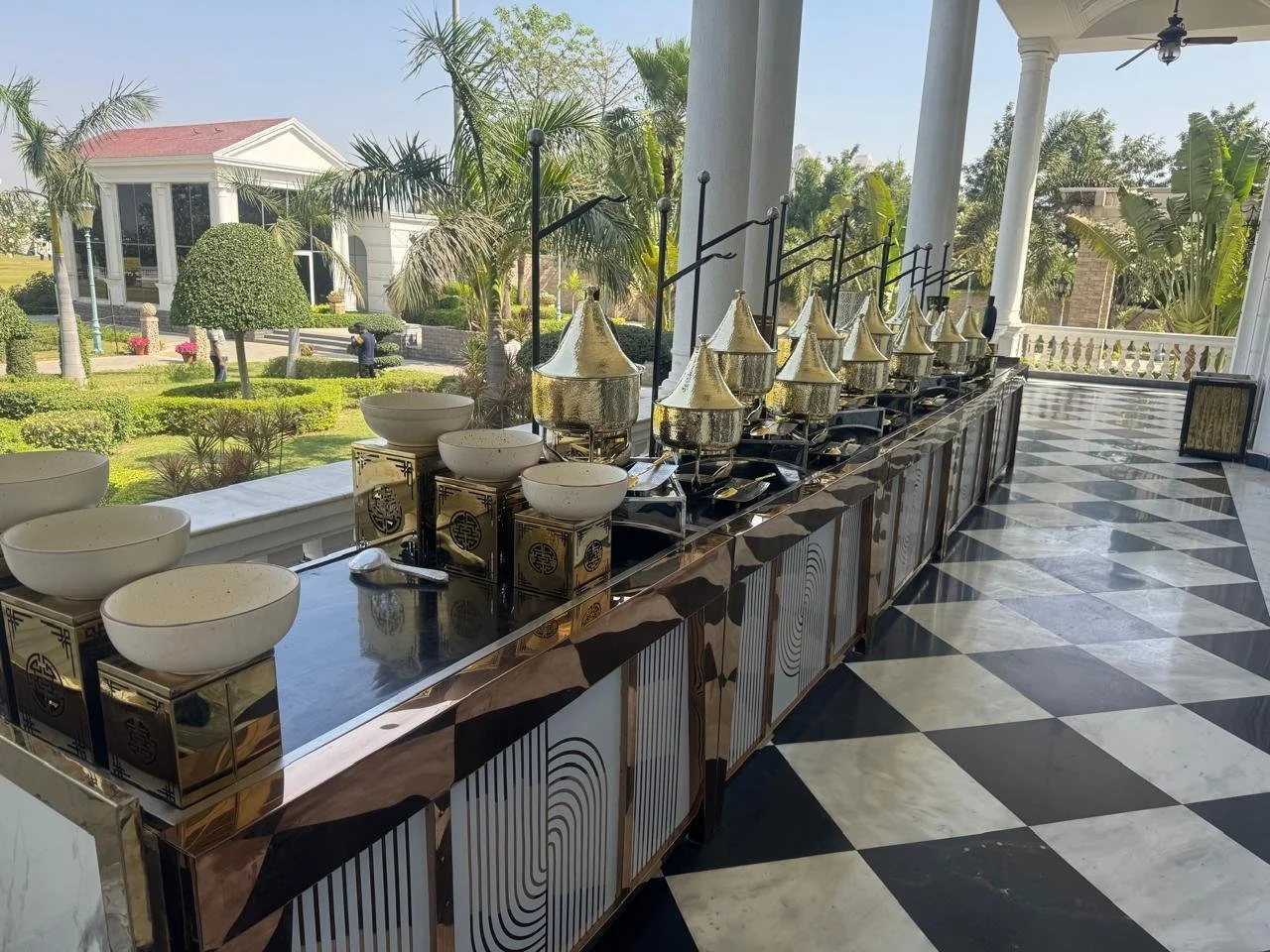 Buffet setup with multiple chafing dishes on a marble counter, white bowls, and a scenic garden view in the background with trees, shrubs, and a white house with a red roof, visible through large windows.