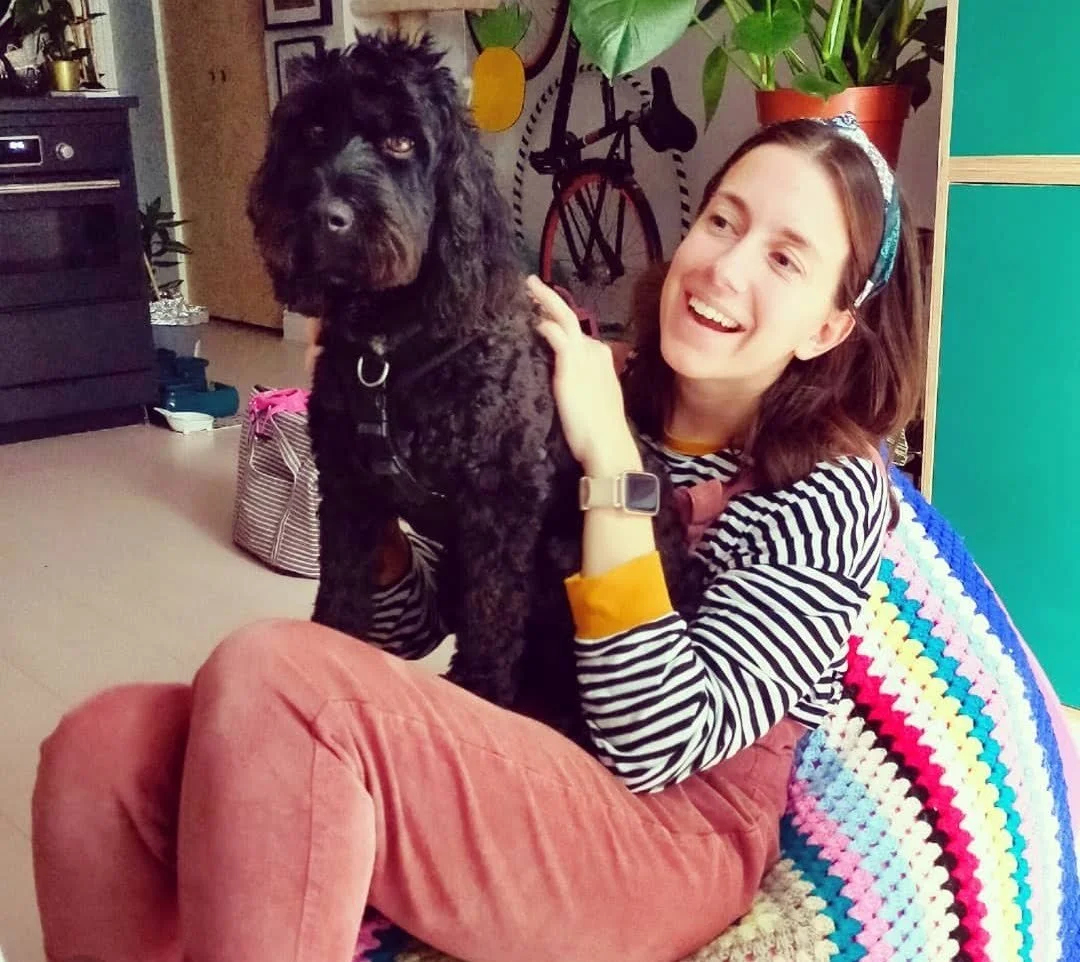 A black cockapoo dog is sat on the lap of a young woman with brown hair wearing dungarees