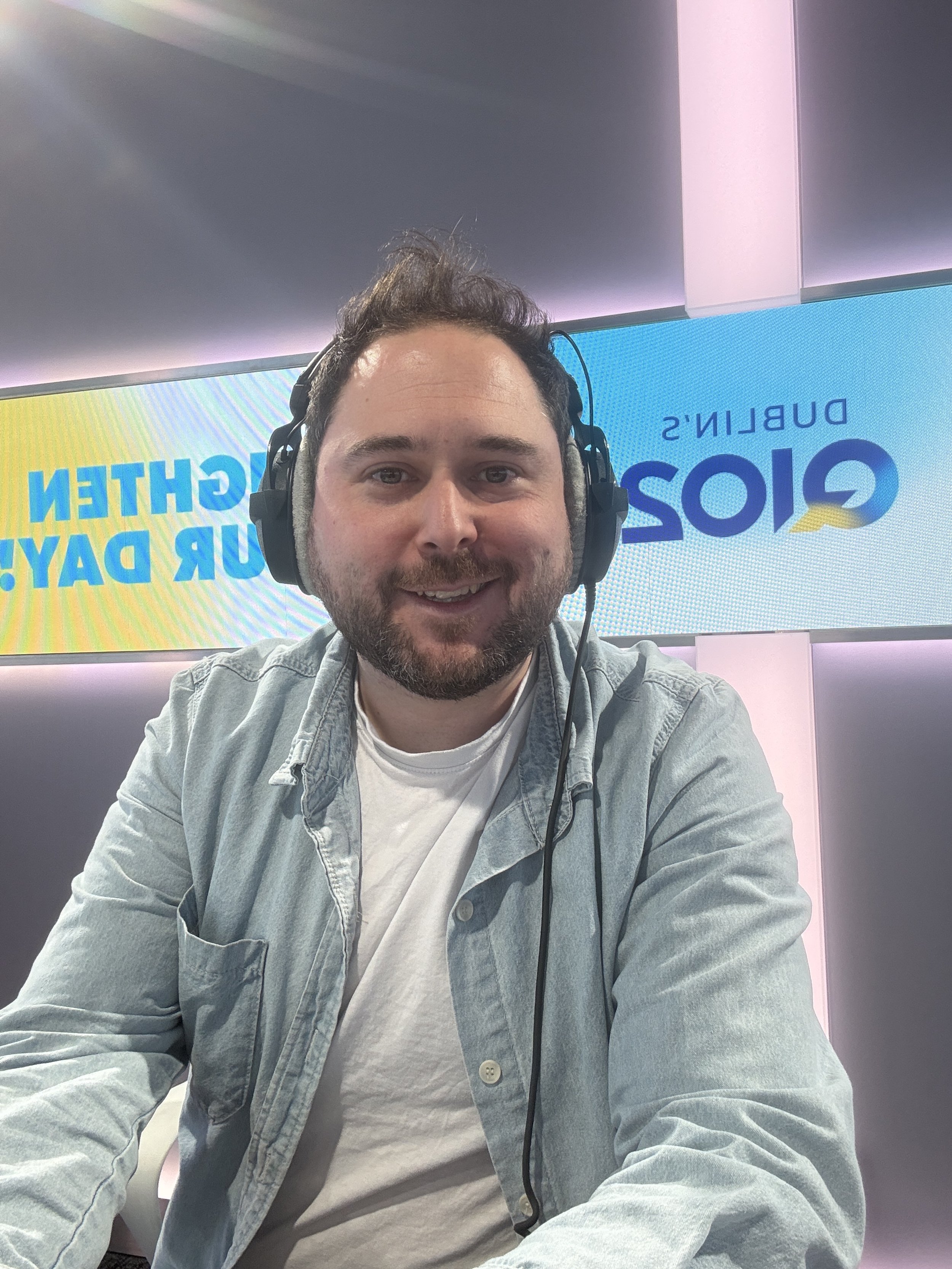 A man with dark hair and a beard wearing headphones, smiling at the camera, seated in front of a screen with the text 'GLOBE' and 'DUBLIN'S' visible.