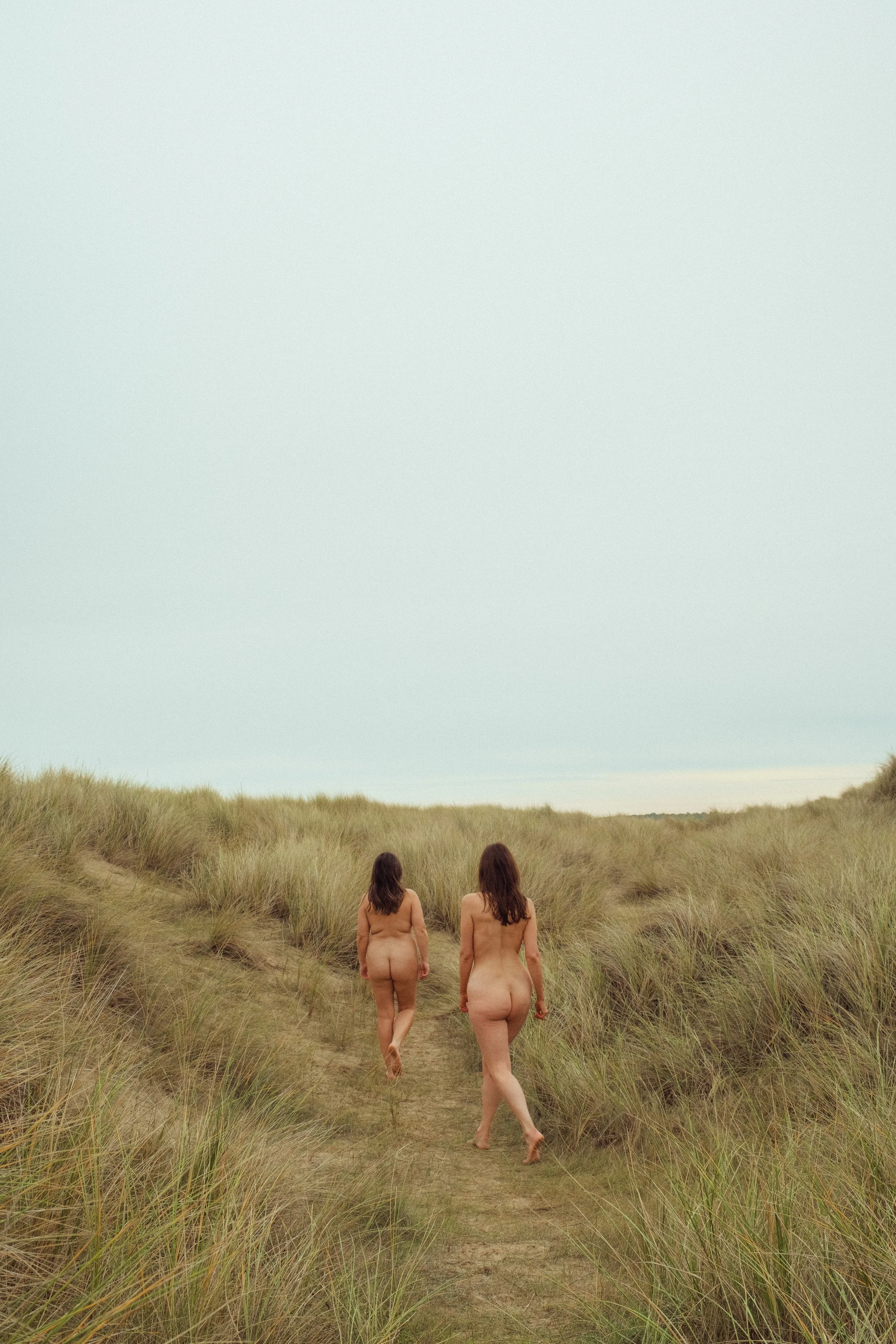 Two nude women walking along a grassy path through sand dunes, away from the camera, under a pale sky.