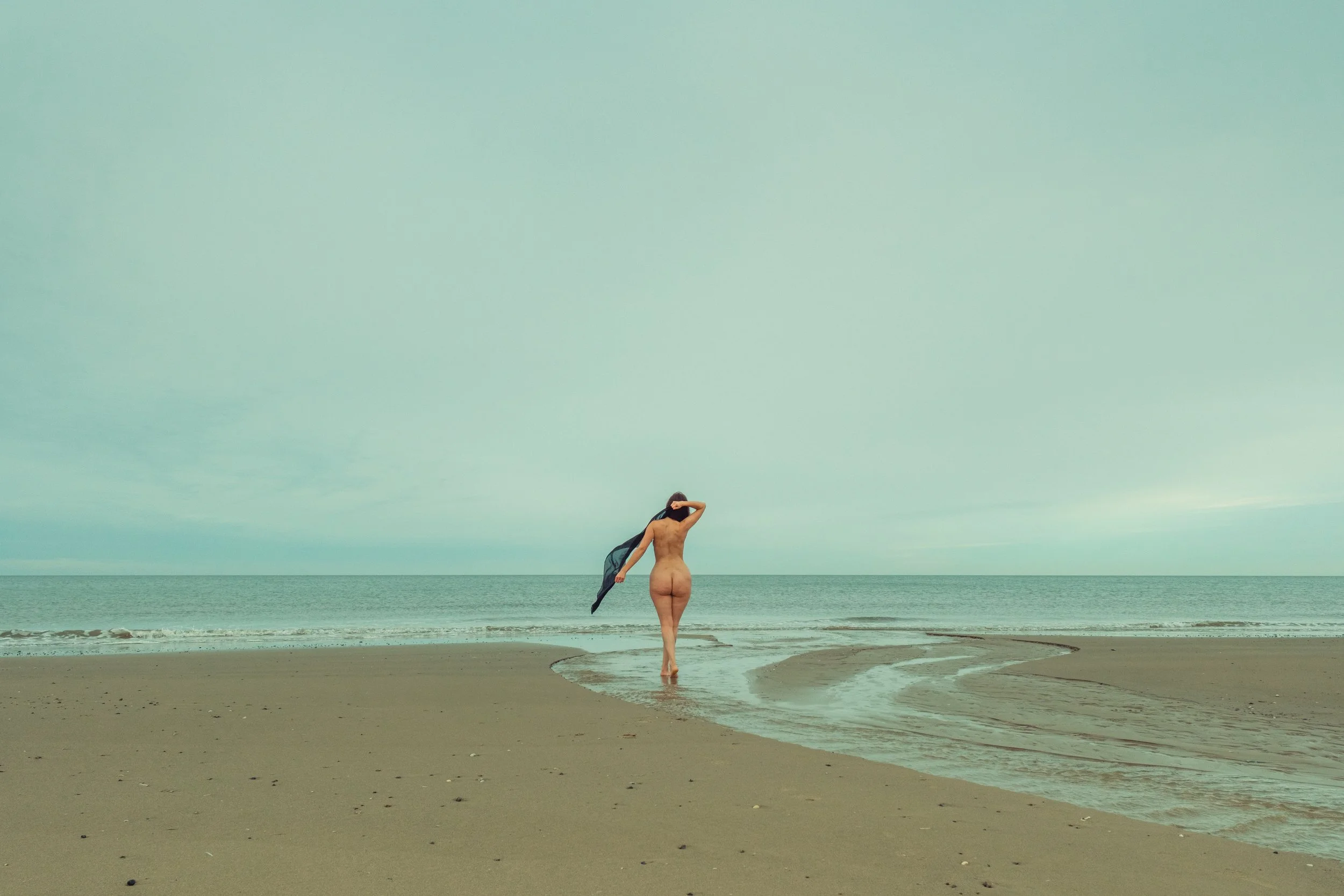 A nude woman walking on a beach with whipped waves, holding a dark scarf, facing away from the camera.