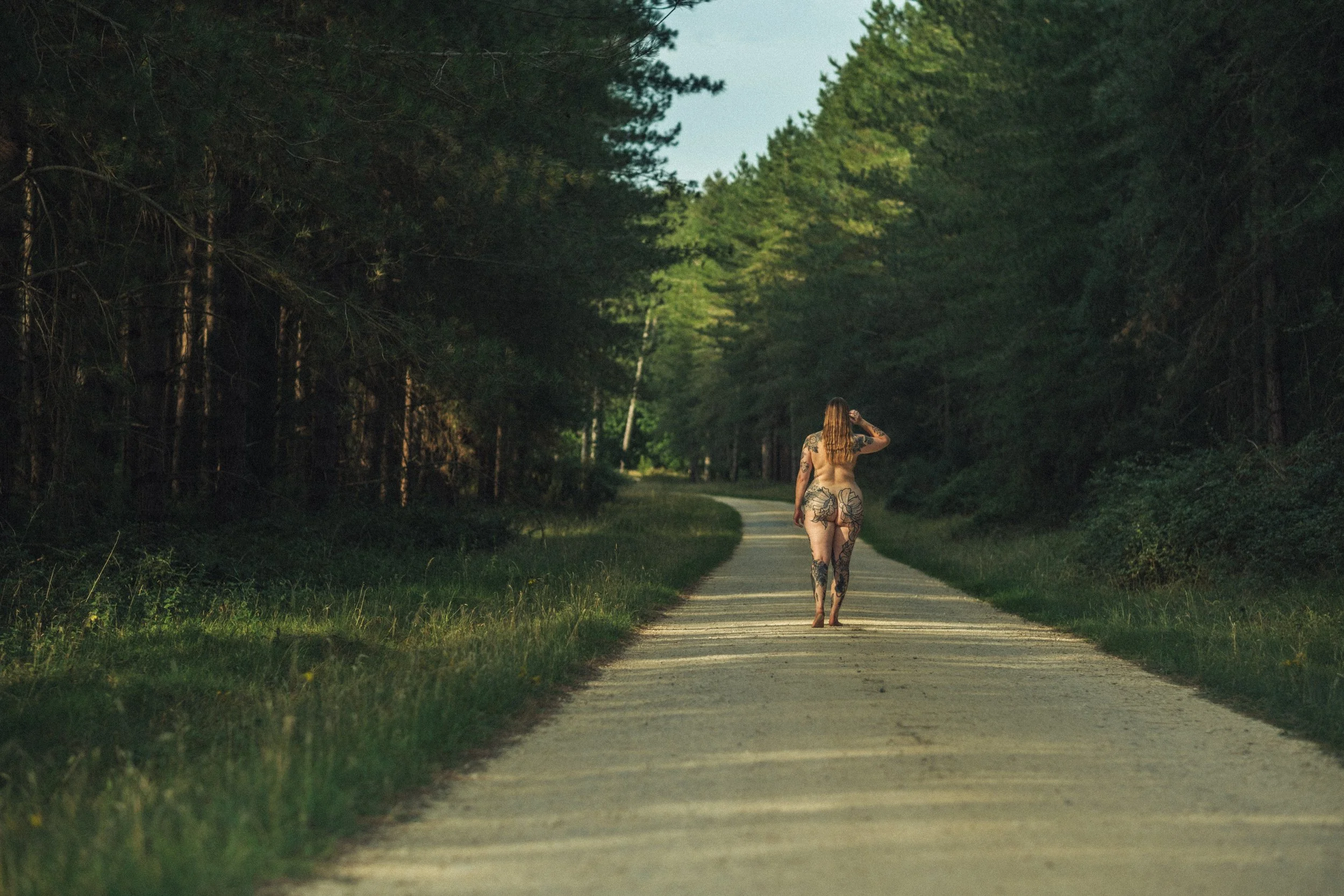 A nude woman with tattoos walking down a dirt forest trail, surrounded by trees and greenery.