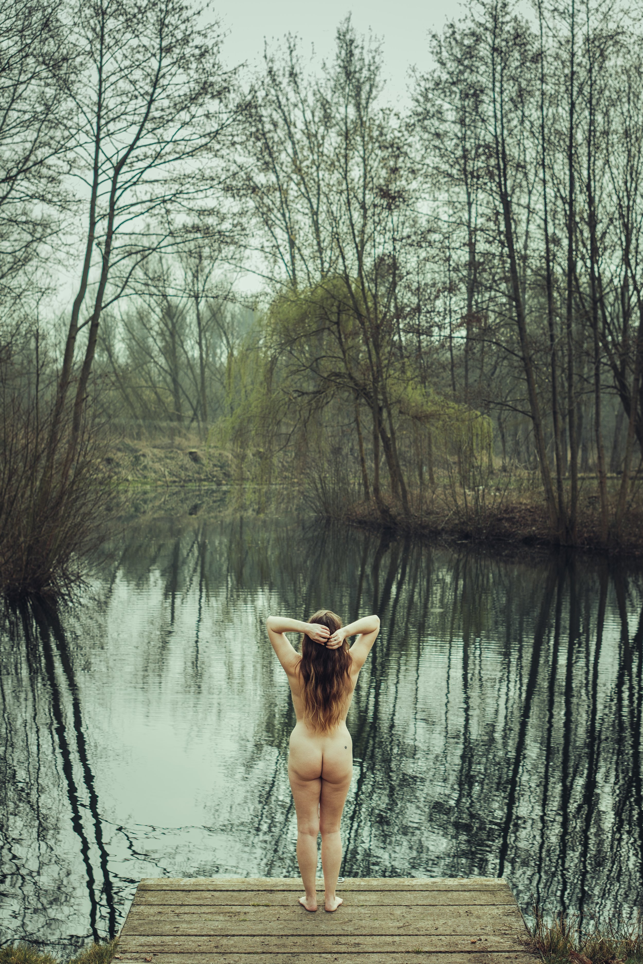 A person standing on a wooden jetty facing a calm lake surrounded by trees, with their back to the camera and hands behind their head.