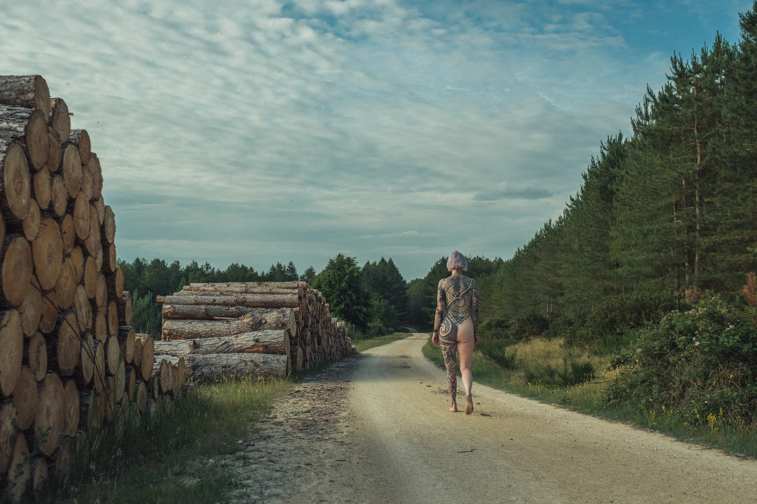 A person with tattoos walking down a rural dirt road, flanked by stacks of logs and a forested area under a partly cloudy sky.