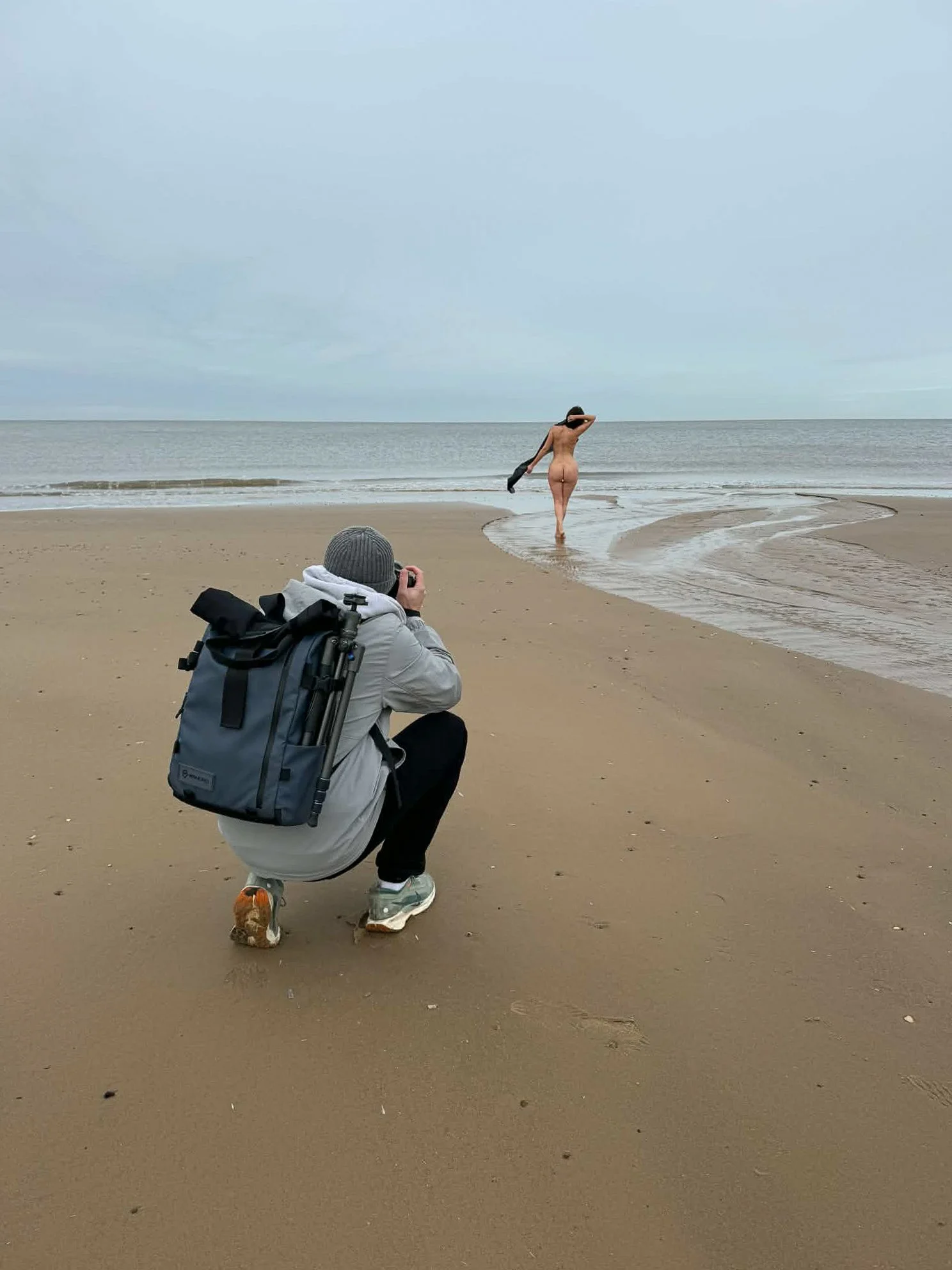 Photographer crouching on a sandy beach taking a picture of a nude woman walking toward the water, surrounded by a cloudy sky and calm ocean.