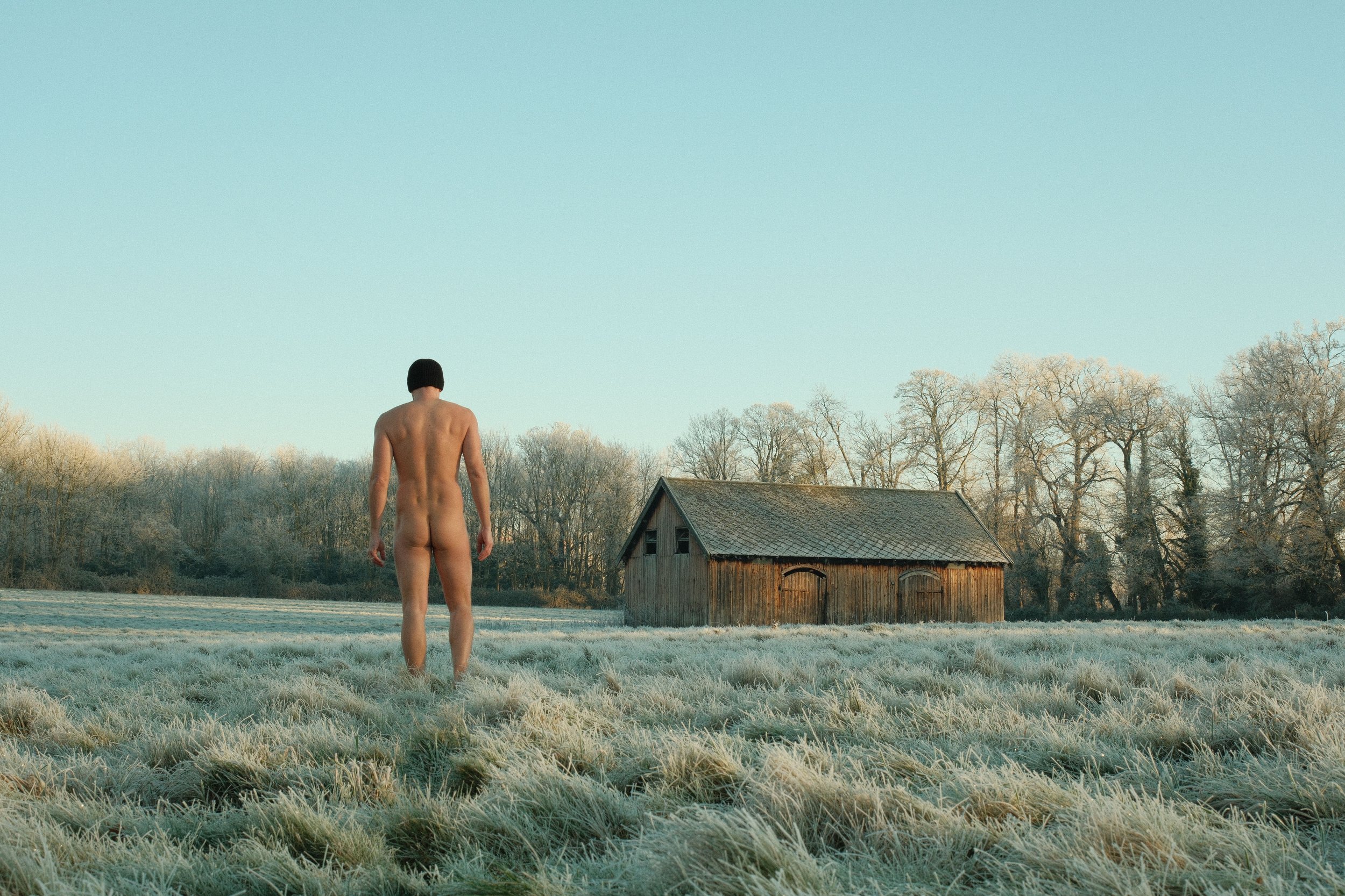 A naked man with a black beanie standing outdoors in a frosty field, facing away from the camera, with a small wooden barn in the background and leafless trees under a clear sky.