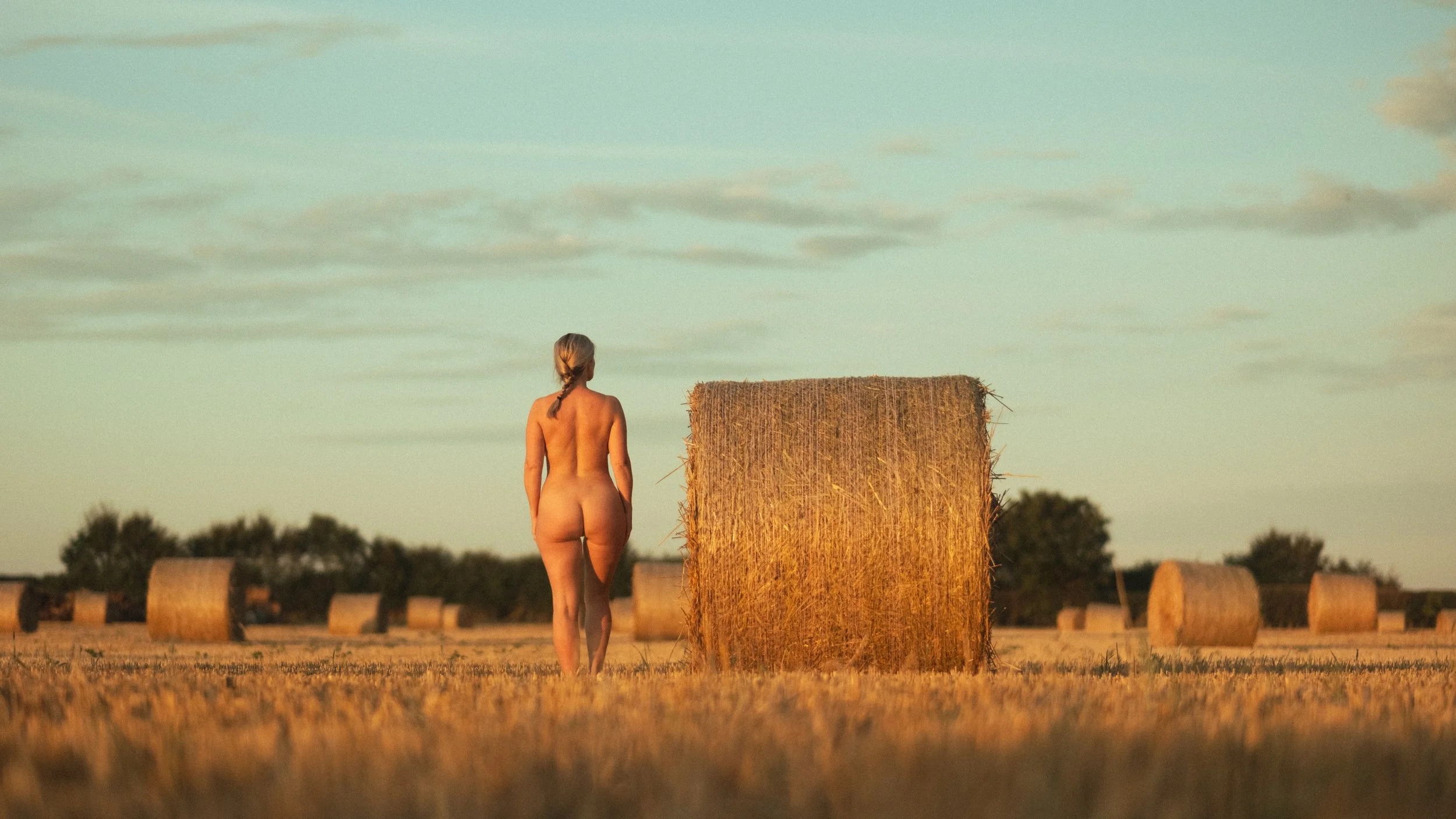 A woman walking nude through a field with hay bales during sunset.